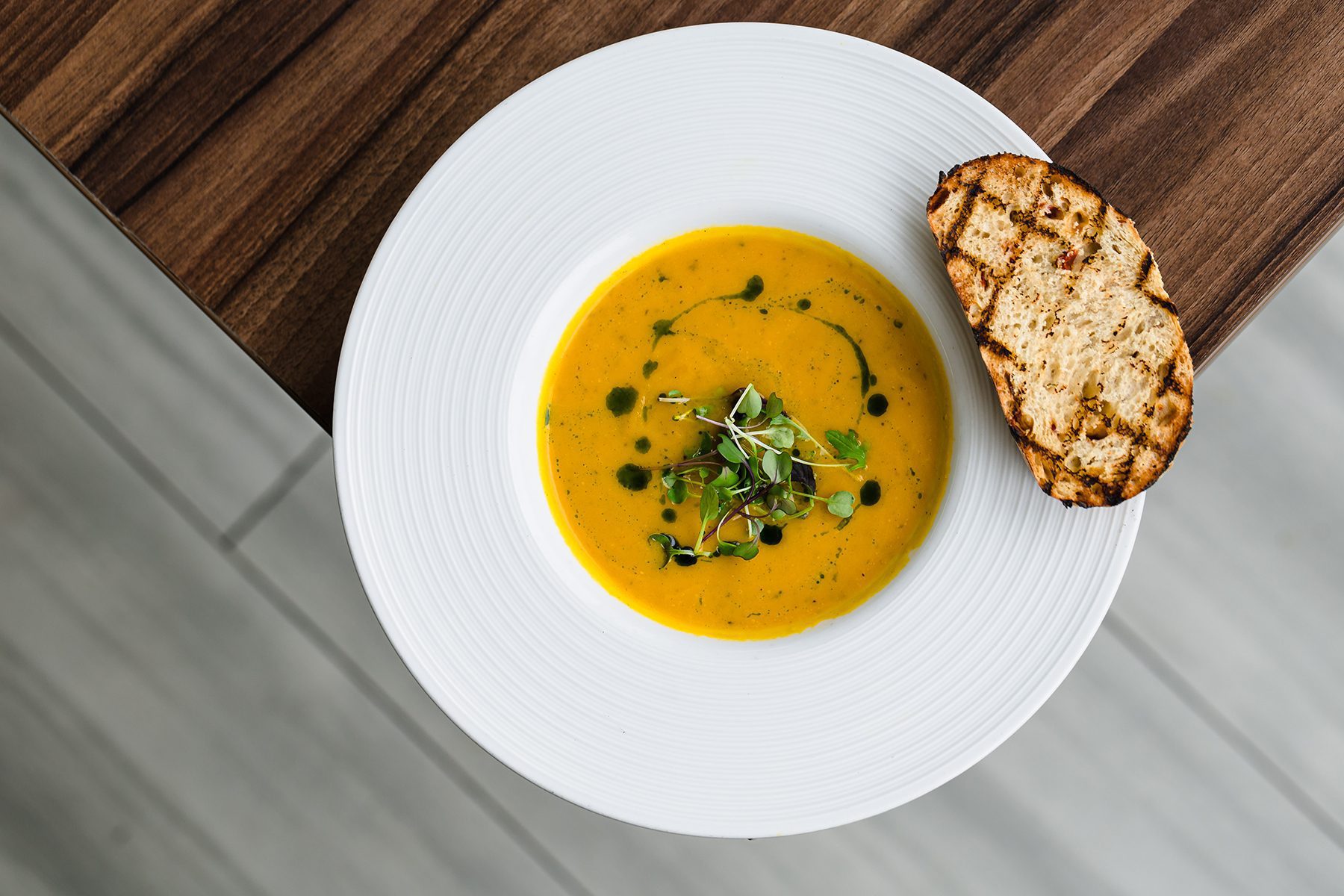 A white plate with pumpkin soup garnished with microgreens and a drizzle of oil, accompanied by a slice of grilled bread, on a wooden table.
