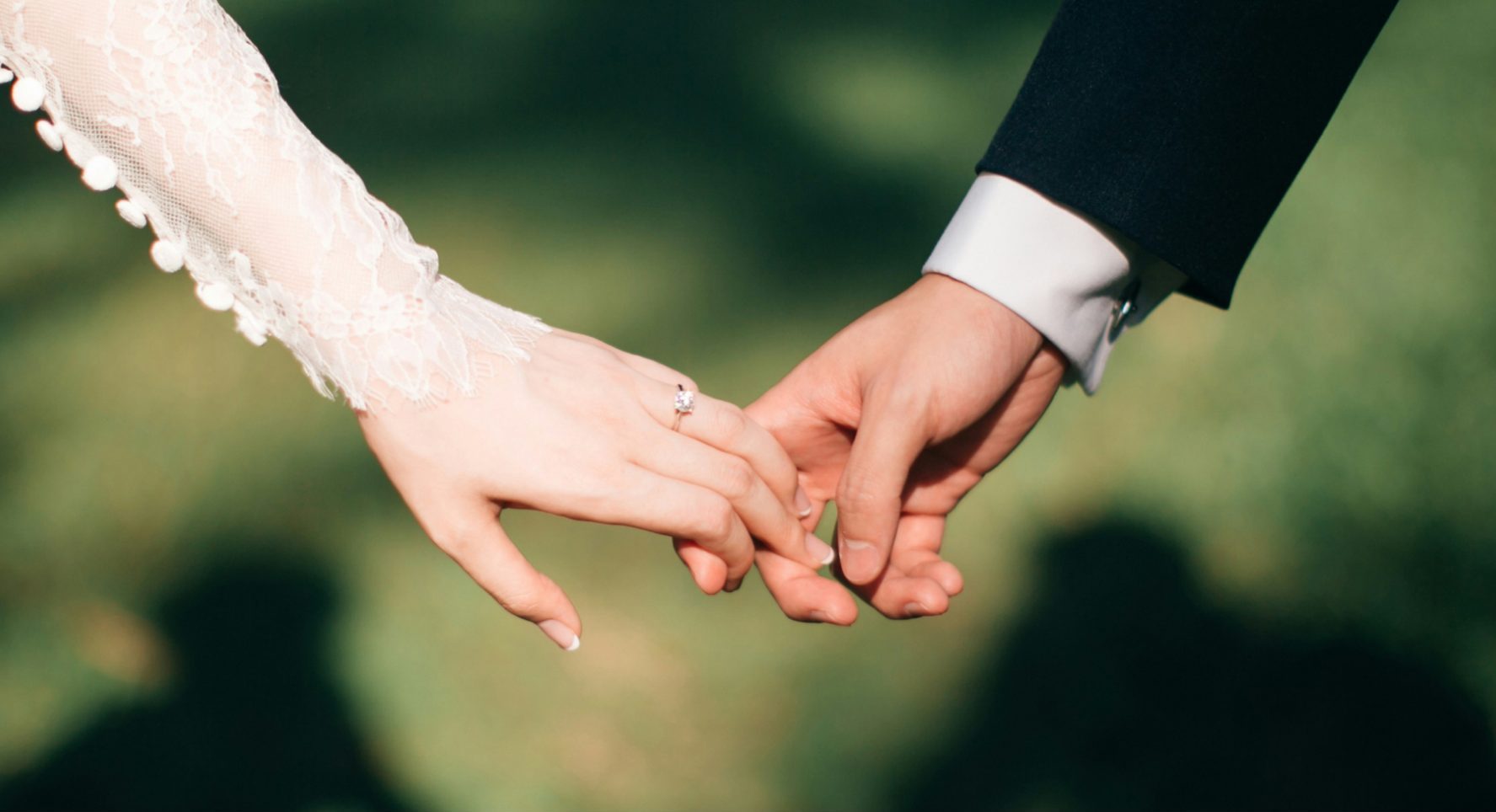 Close-up of a couple holding hands. The woman's hand has a lace sleeve, while the man's hand is in a dark suit. The background is a blurred green outdoor scene.