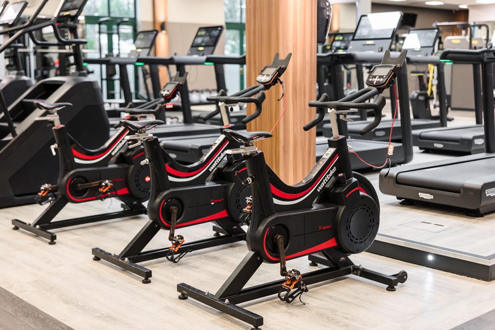 Three stationary exercise bikes are lined up in a gym, with treadmills visible in the background.