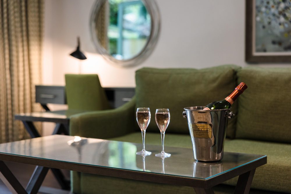 A bottle of champagne in an ice bucket and two filled glasses sit on a glass coffee table in a modern living room.