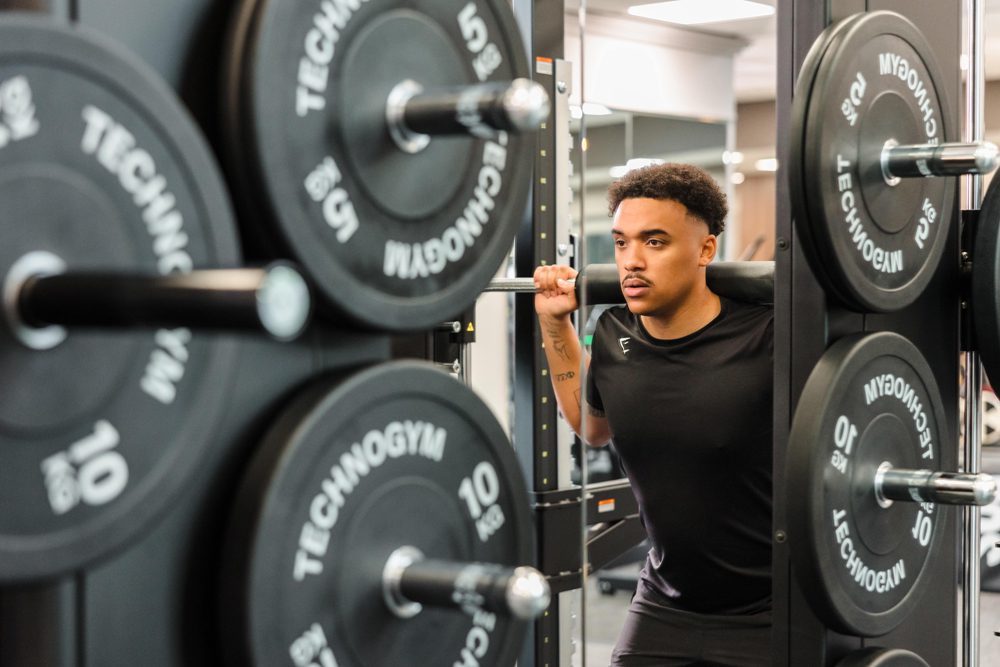 A person wearing a black shirt performs a barbell squat in a gym with weight plates labeled 
