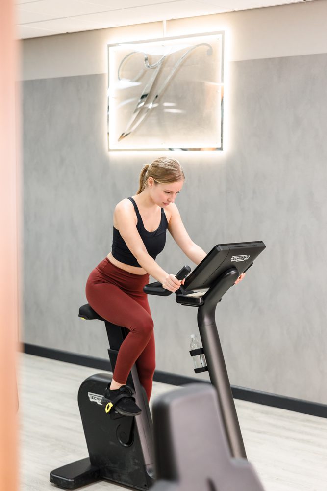 A woman in workout clothes uses a stationary exercise bike in a modern Hotel Gym South Wales with a gray wall and sign behind her.