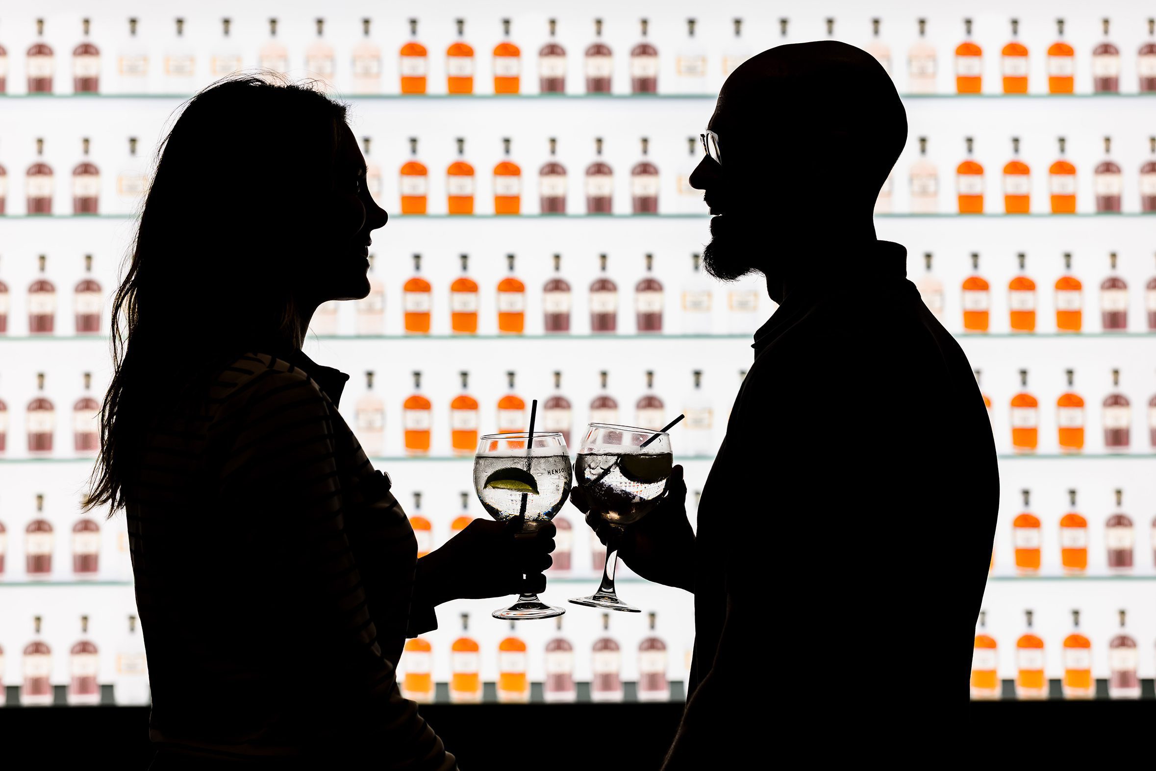 Silhouettes of two people holding drinks, standing by shelves of bottles, discuss hotel room bookings.