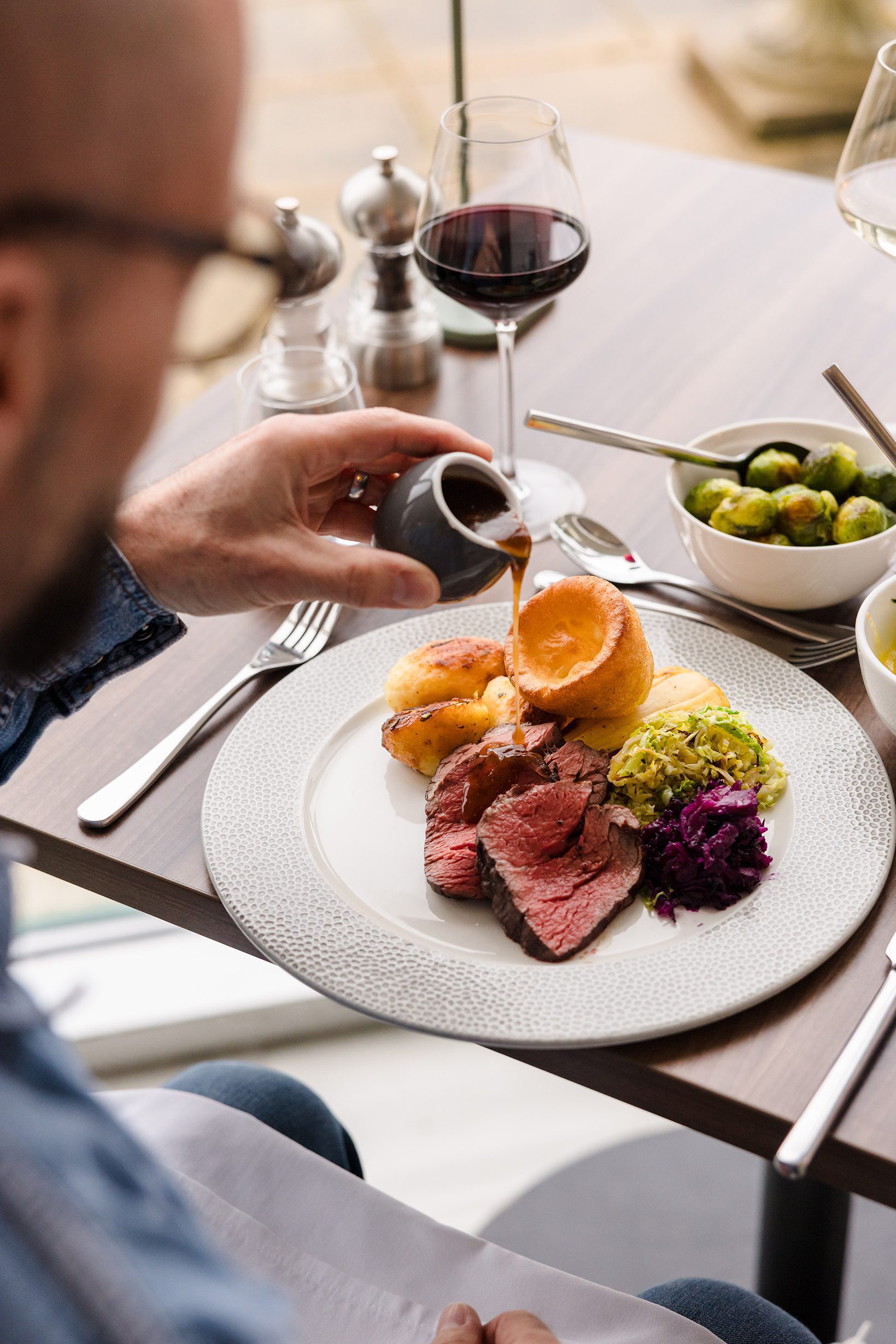A person enjoys dining, pouring gravy over roast beef, Yorkshire pudding, potatoes, and veggies next to a glass of red wine.