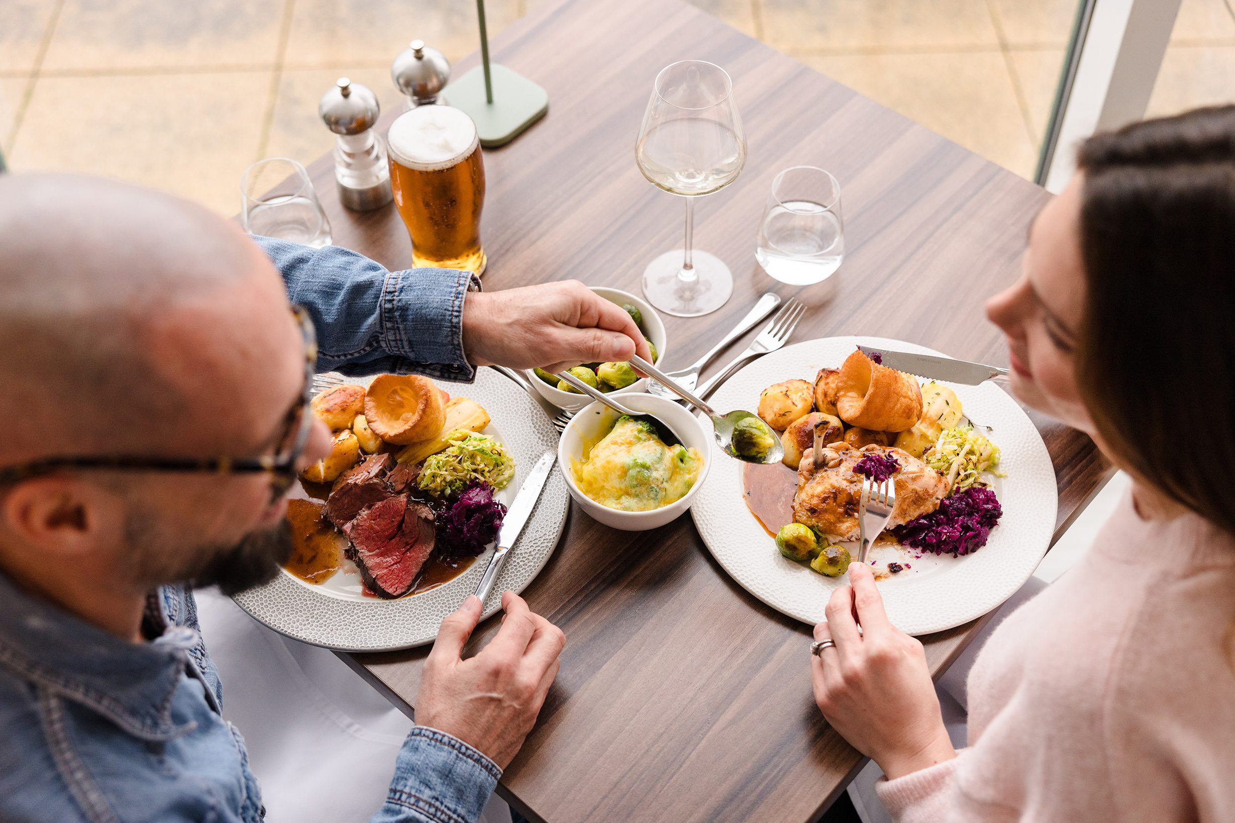 Two people enjoy a Sunday lunch at a restaurant table with roast meat, vegetables, drinks, and cutlery visible.