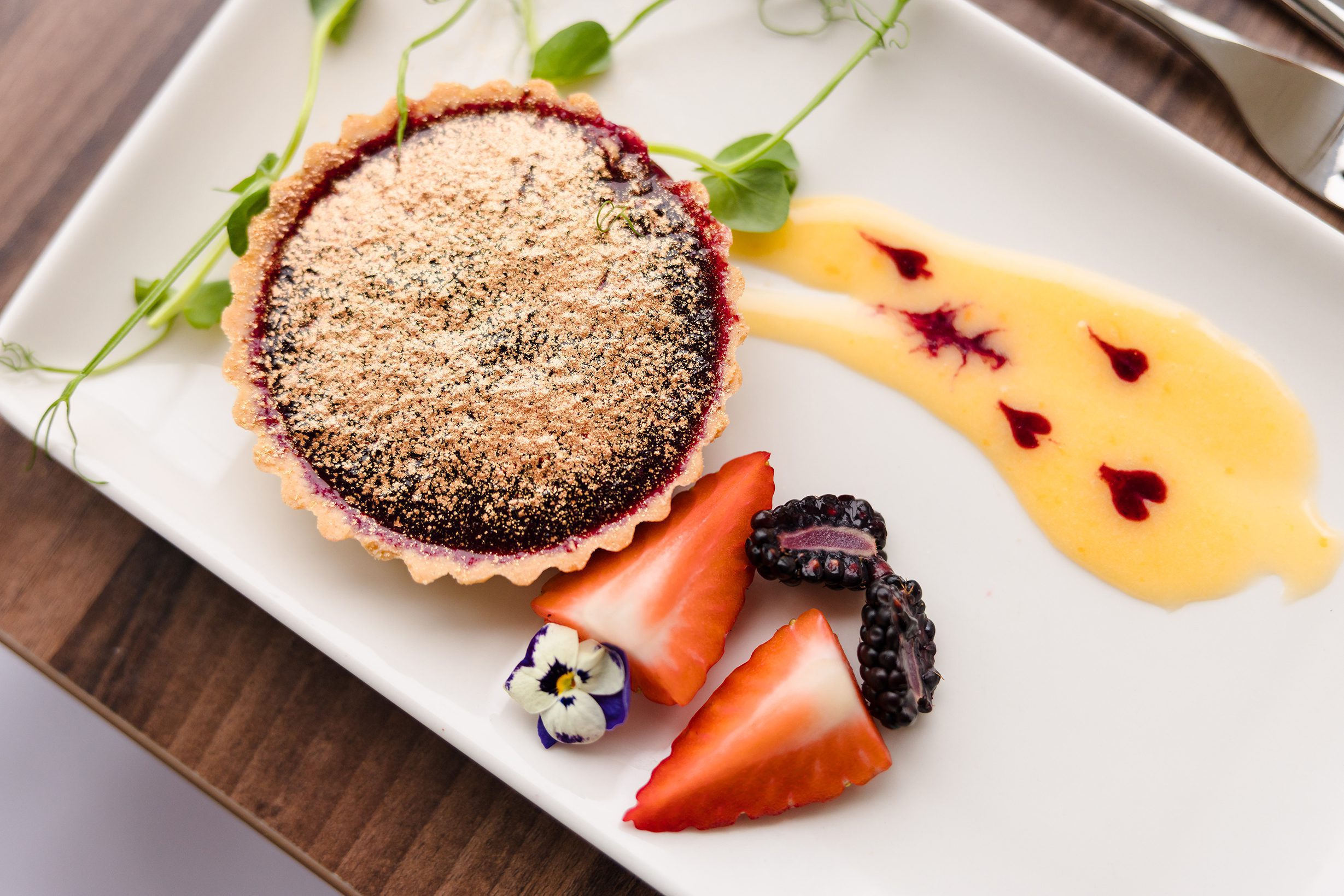 A fruit tart on a white plate, perfect for sunday lunch, with strawberries, blackberries, edible flowers, and microgreens.