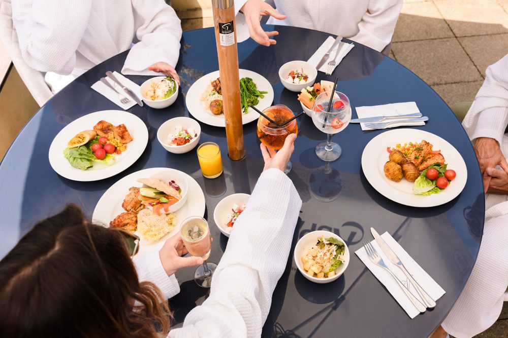 Four people in white robes enjoy a meal on a round outdoor table during relaxing Spa Breaks South Wales.