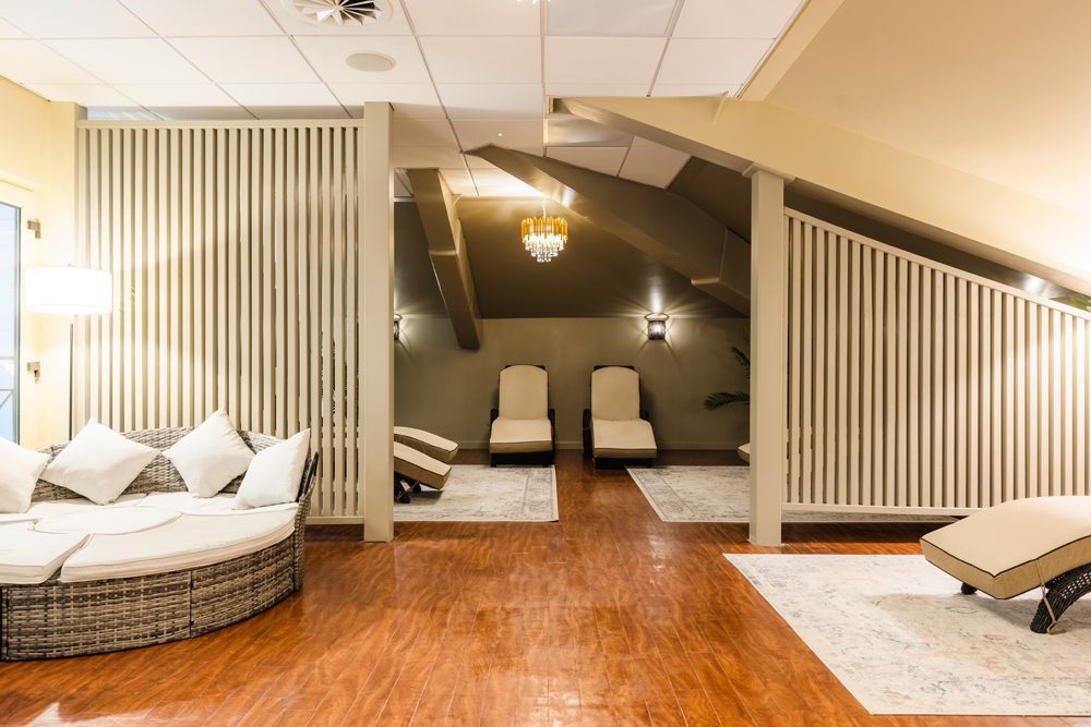 A modern spa lounge area with wicker furniture, lounge chairs, wood flooring, and soft lighting under a sloped ceiling.