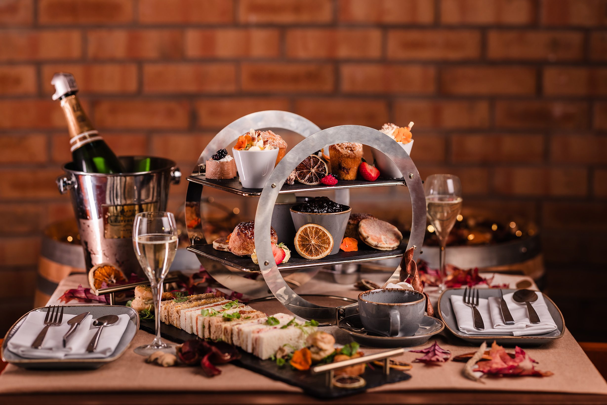 A table set for afternoon tea with tiered stands of pastries, a champagne bottle in a bucket, and elegant place settings.