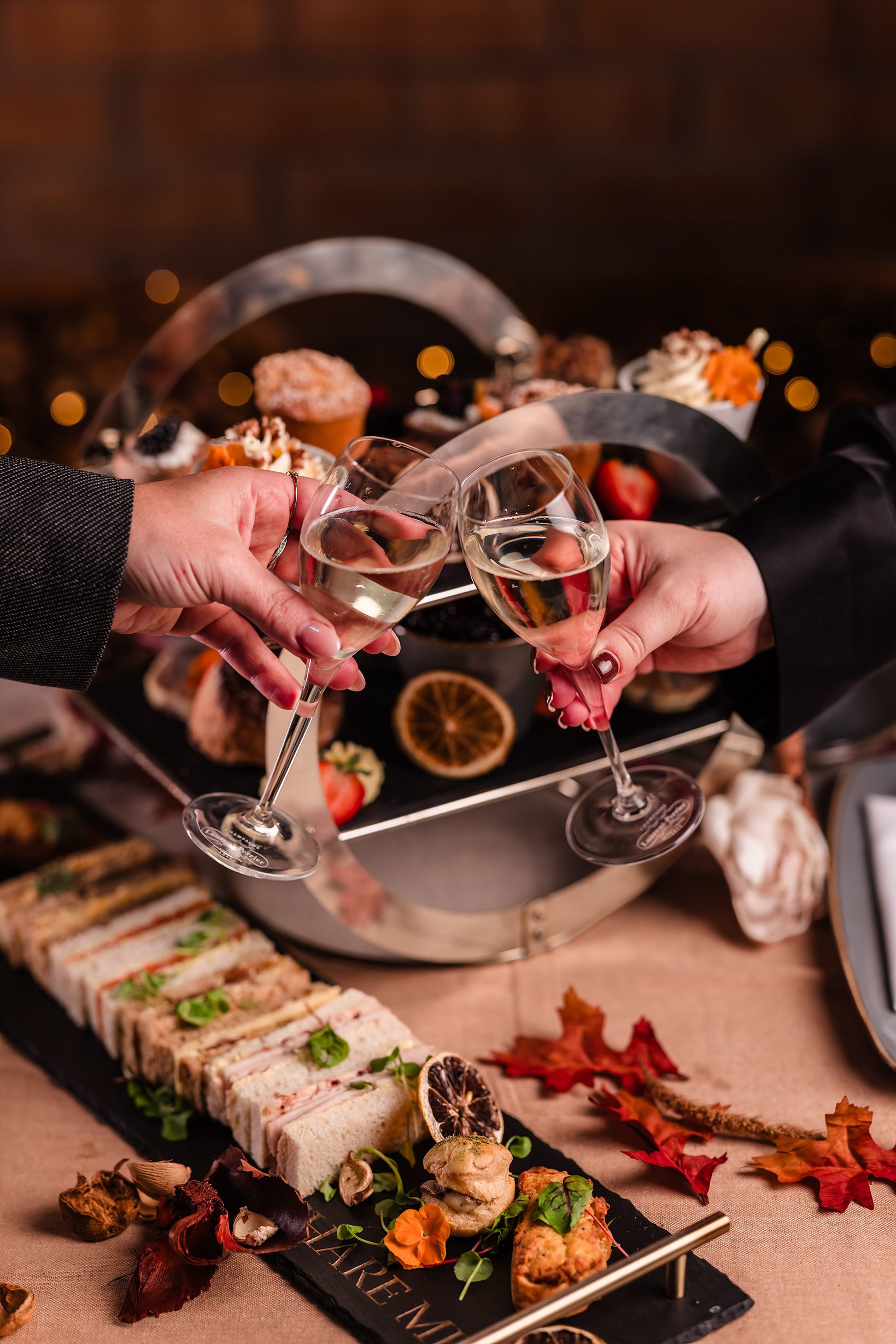 Two people enjoy afternoon tea, clinking champagne glasses over a table of sandwiches, pastries, and autumn decor.