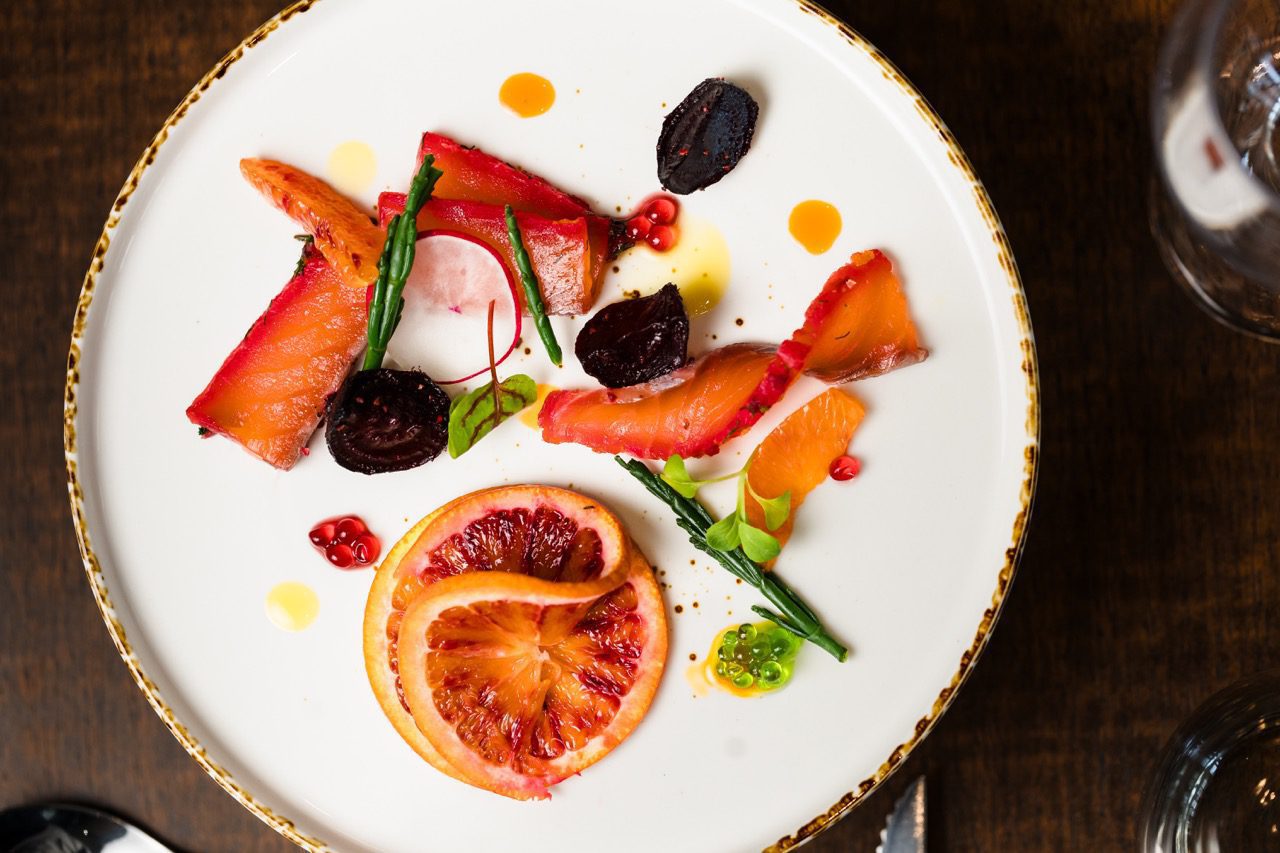 Plate of gourmet food with slices of cured fish, blood orange, beets, and green garnishes on a white dish.