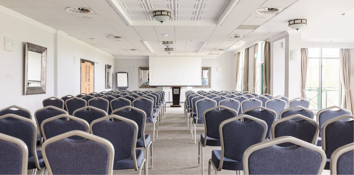 Empty conference room with rows of blue chairs facing a projector screen in a bright setting.