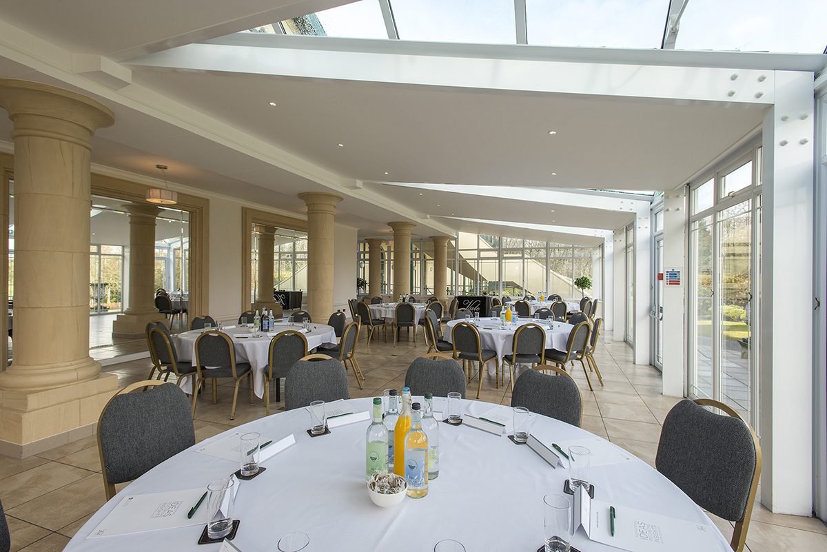 A spacious event room with several round tables covered with white tablecloths, surrounded by chairs. The room has large windows and columns, with bottles and glasses on the tables.