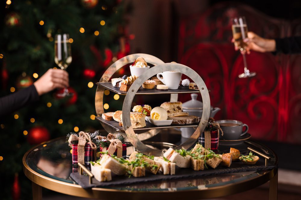 A tiered tray with assorted pastries and sandwiches on a table, with two people holding champagne glasses in the background.