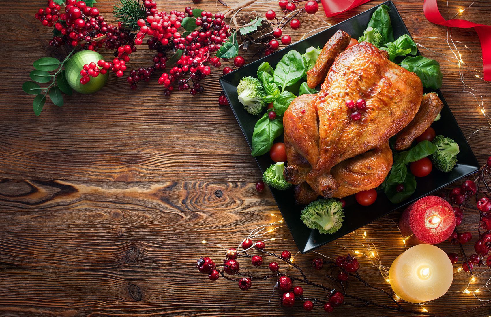 A roasted chicken on a black platter surrounded by greens and tomatoes. The table is decorated with red berries, a candle, and Christmas ornaments.