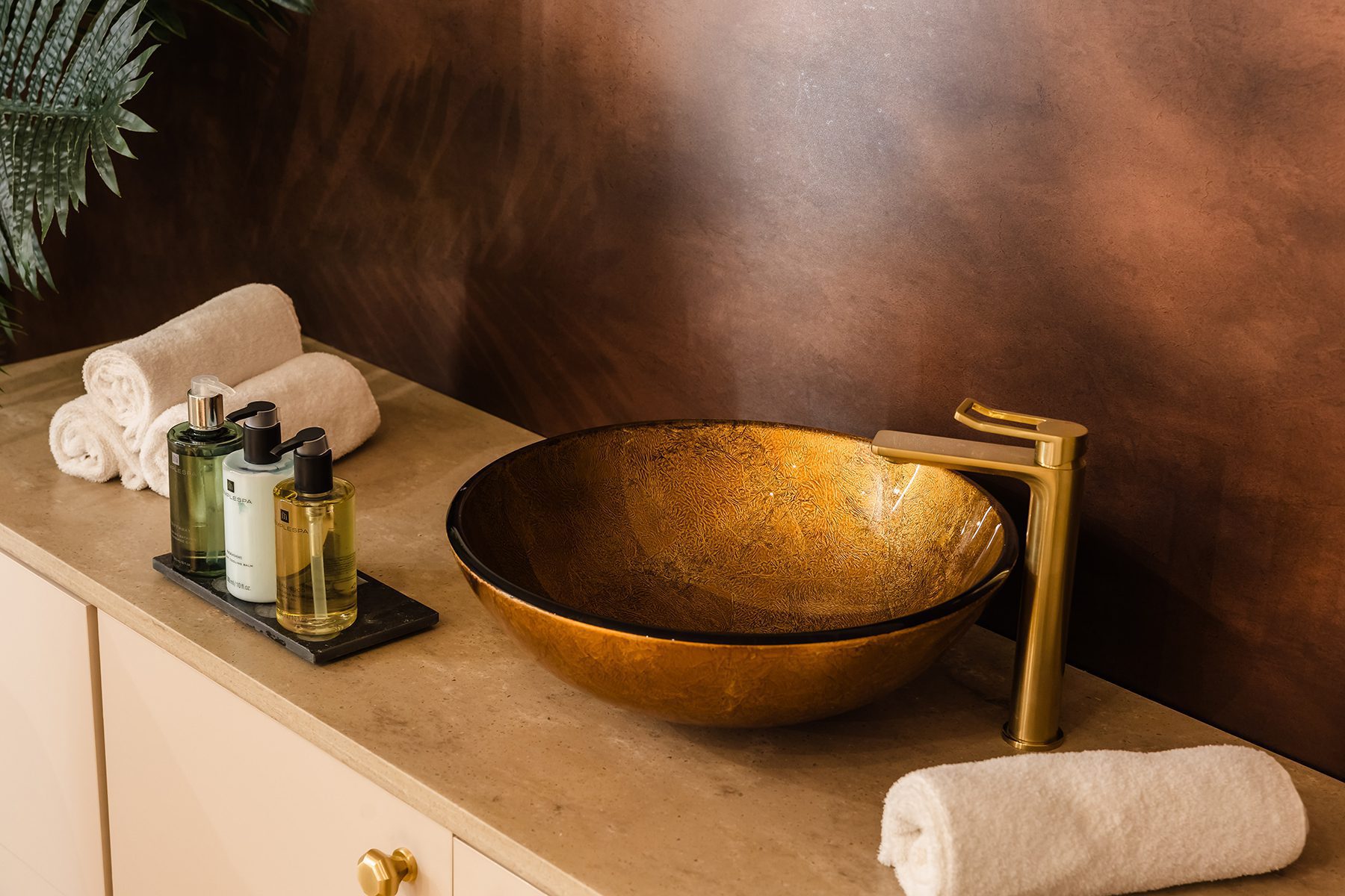 gold vessel sink, matching faucet, rolled towels, and toiletries on a beige countertop against a brown wall.