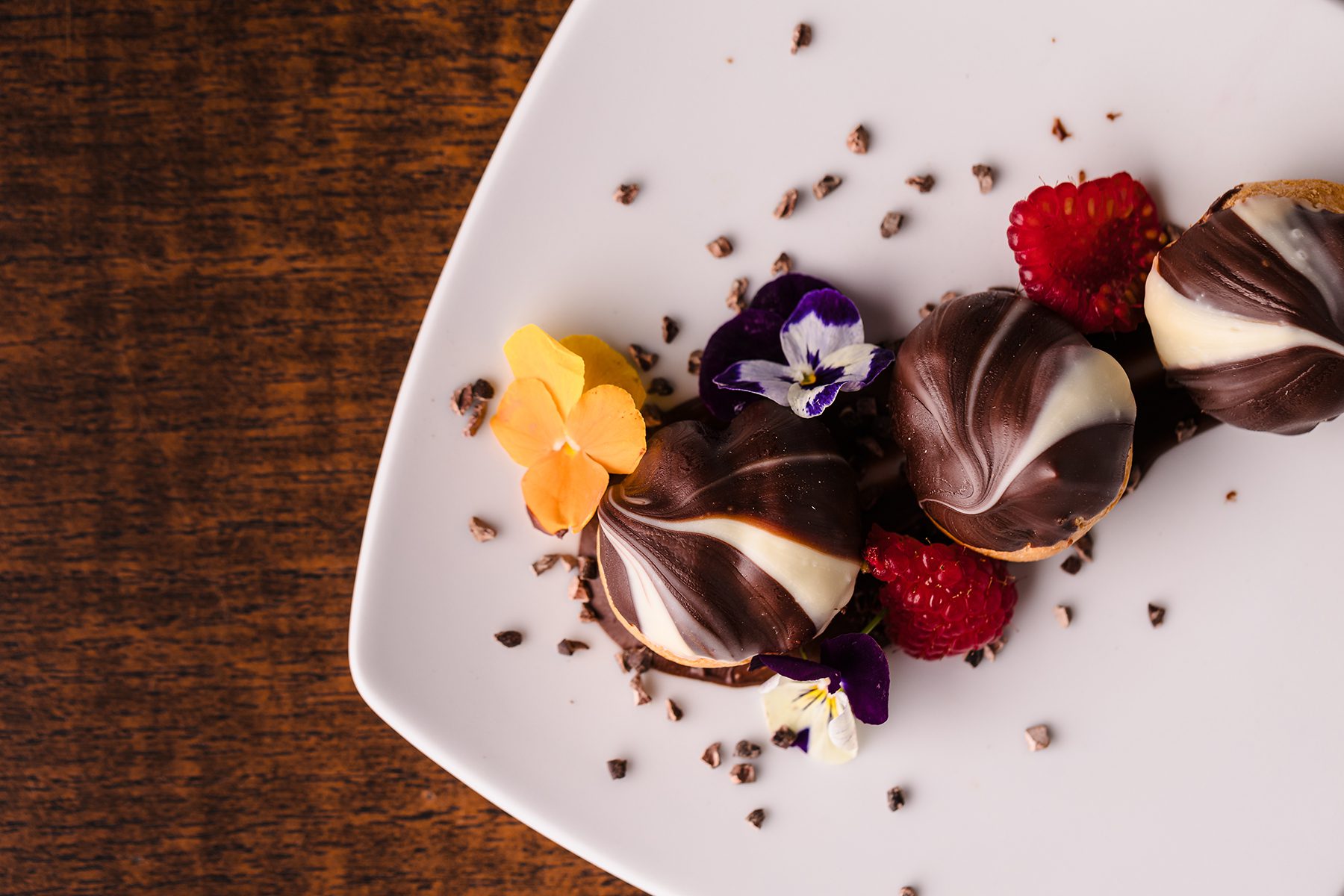 Chocolate-covered pastries with white chocolate swirls, garnished with raspberries, pansies, and edible flowers on a white plate against a wooden background.