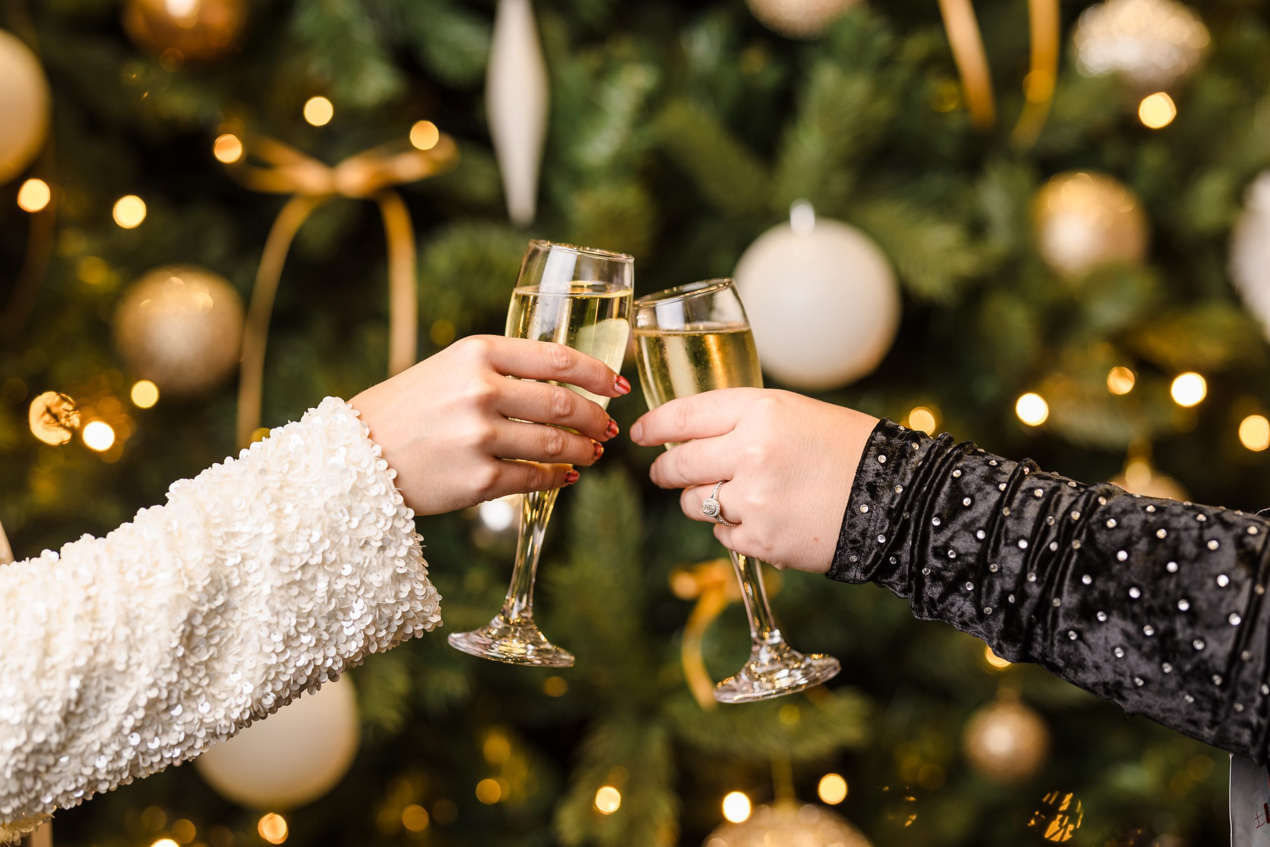 Two people clink champagne glasses in front of a decorated Christmas tree with gold and white baubles.