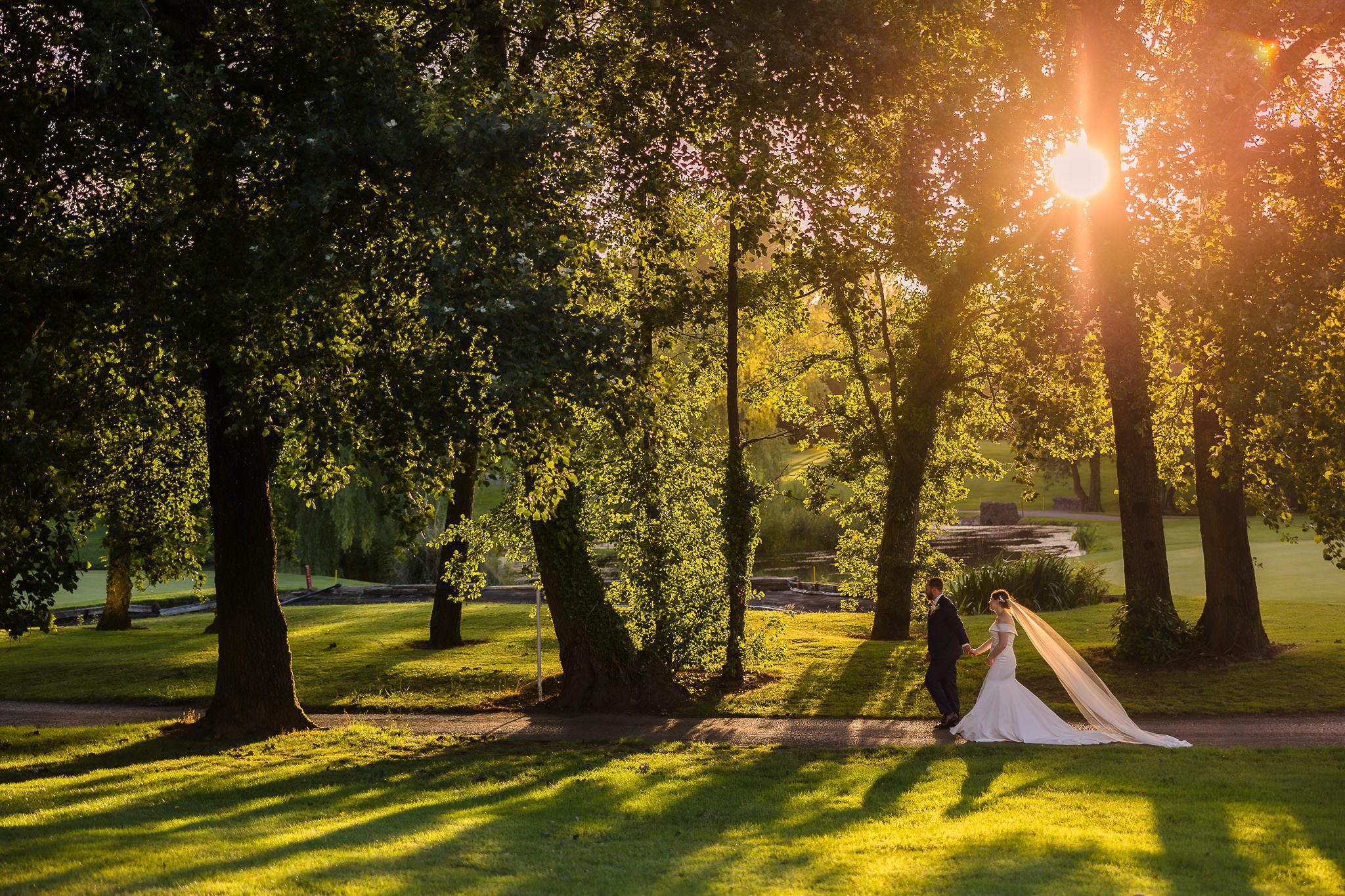 A couple walks hand in hand in a sunlit park. The bride's veil flows behind her as sunlight filters through the trees, casting long shadows on the grass.