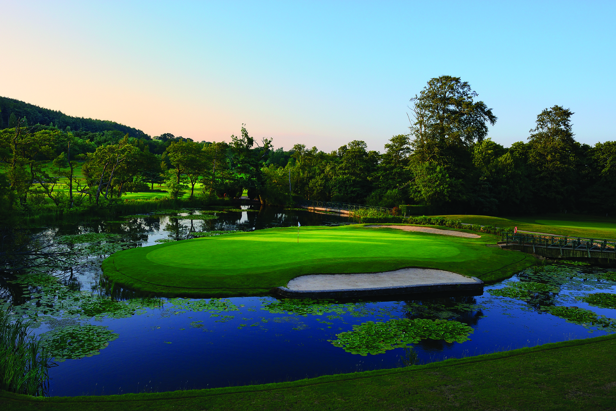 A golf course with a green surrounded by water and trees under a clear blue sky.