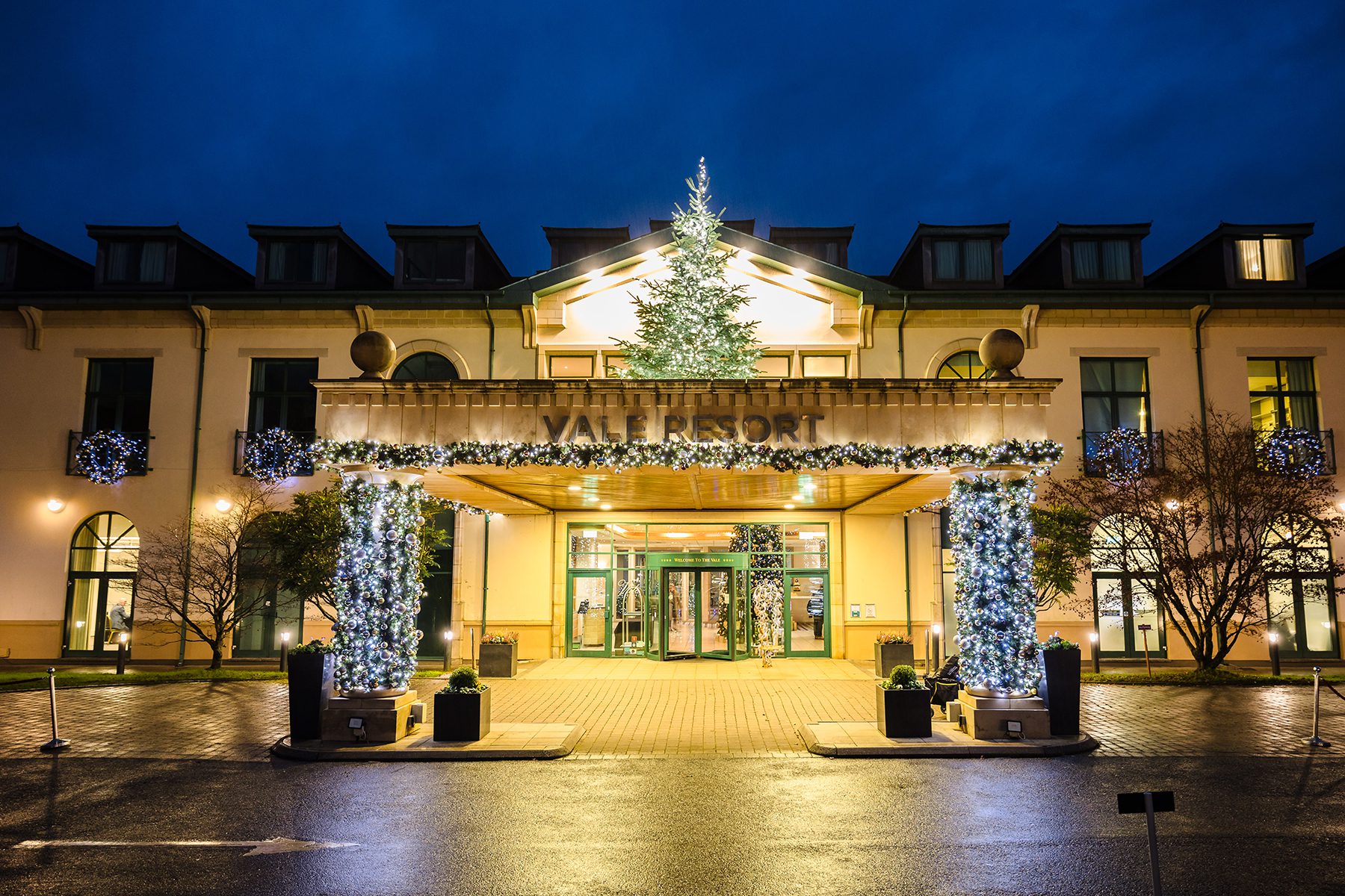 Hotel entrance decorated with Christmas lights and a Christmas tree at night, with wet pavement in front.