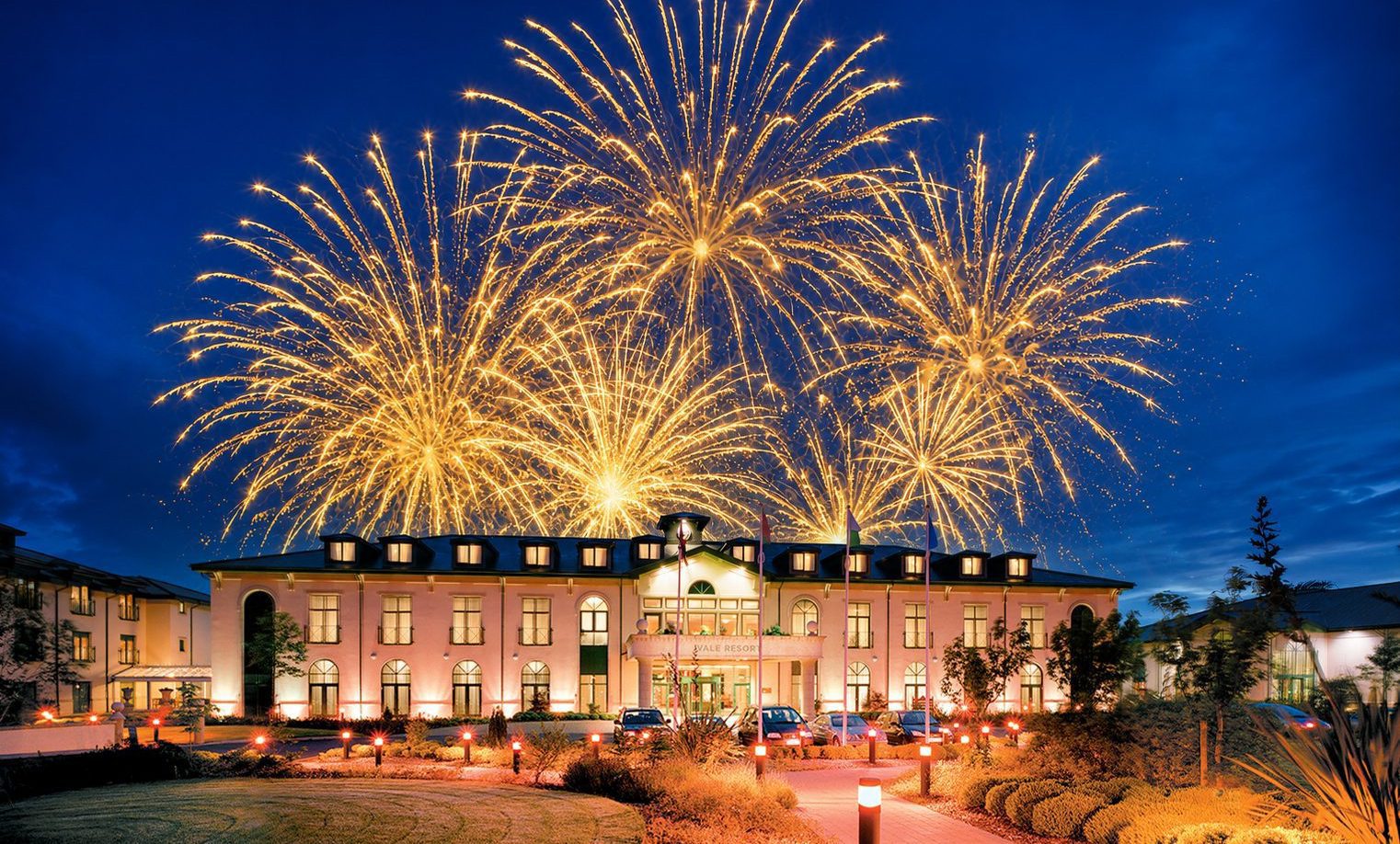 Large gold fireworks explode in the night sky above a brightly lit, grand hotel building with a manicured drive.