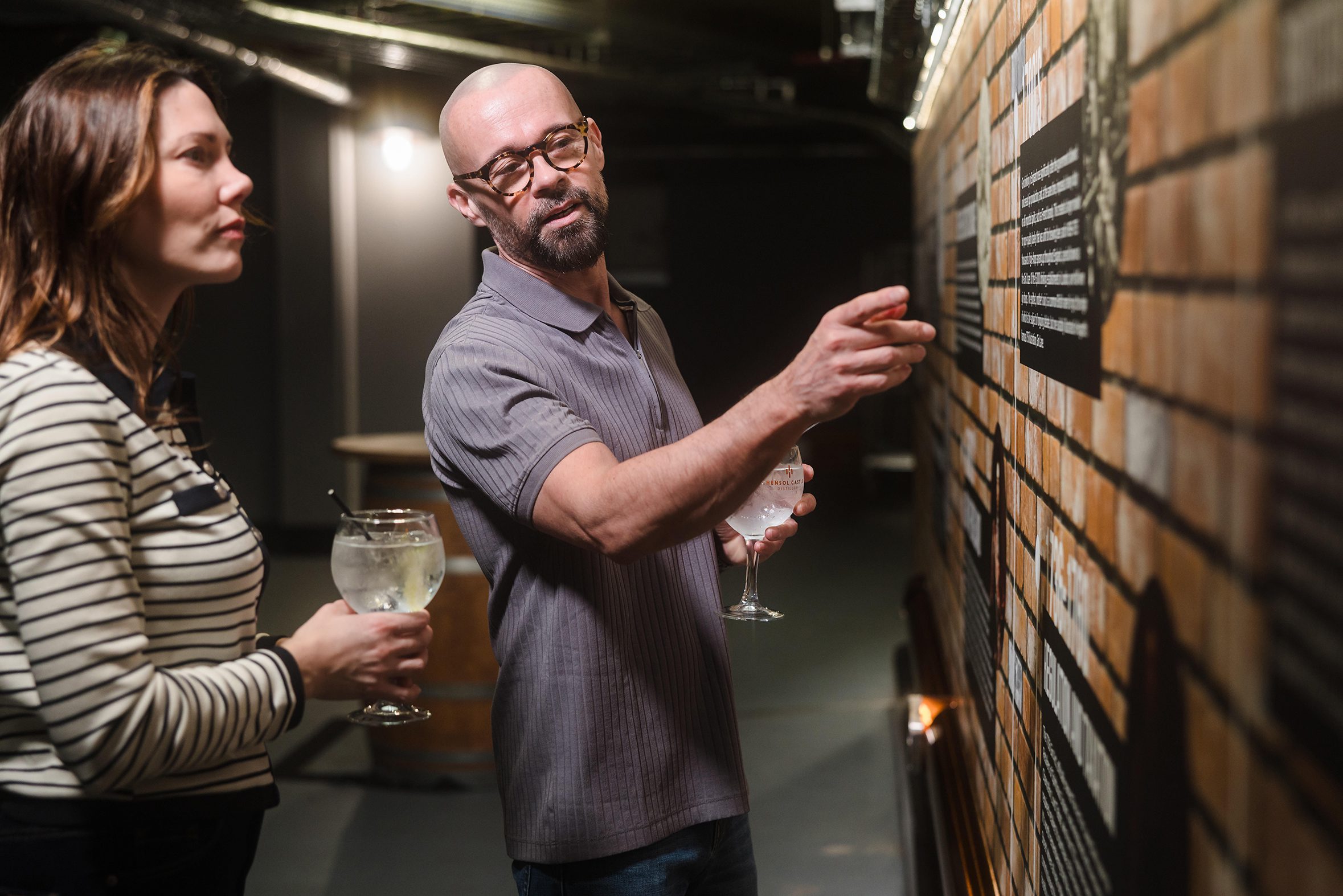 Two people discuss upcoming concerts Cardiff as they examine text on a wall display in a dimly lit indoor setting.