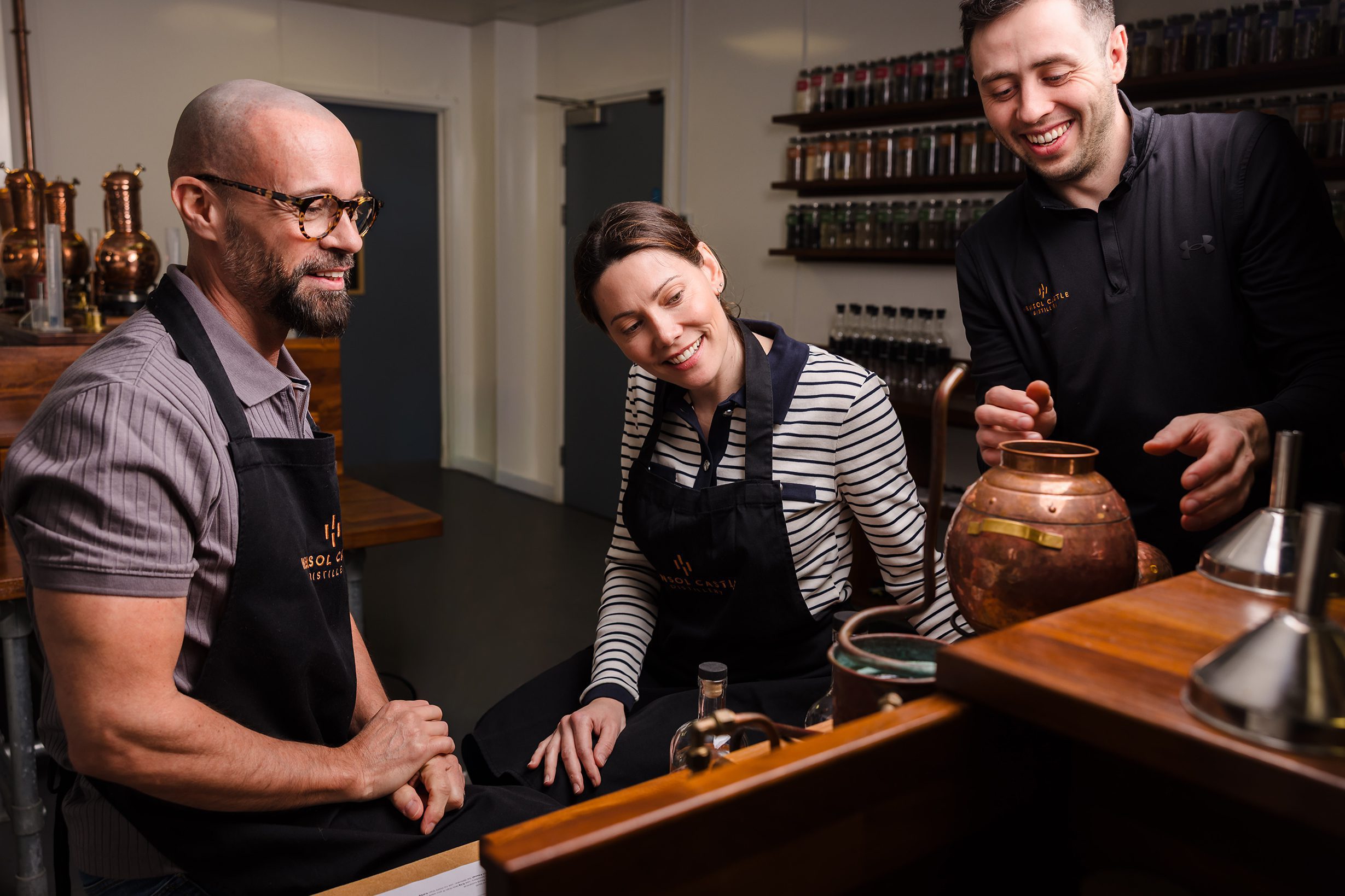 Three people in aprons smile and work together around a copper pot, chatting about upcoming concerts in Cardiff.