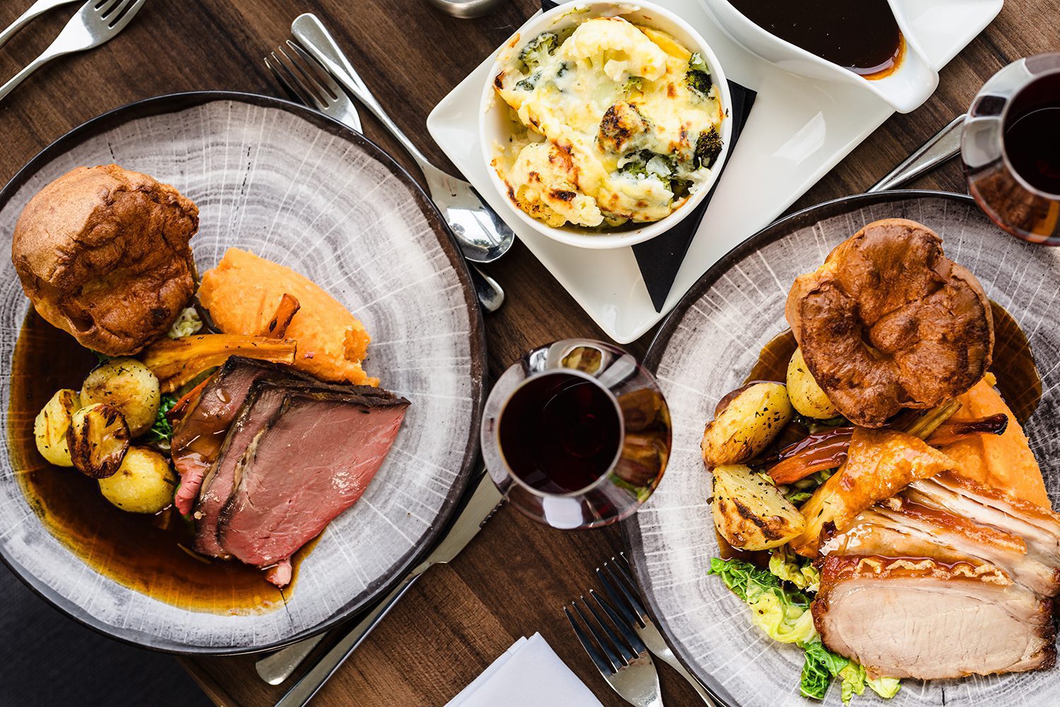 Two plates of roast meat with potatoes, vegetables, and Yorkshire pudding, served with wine and a side dish.