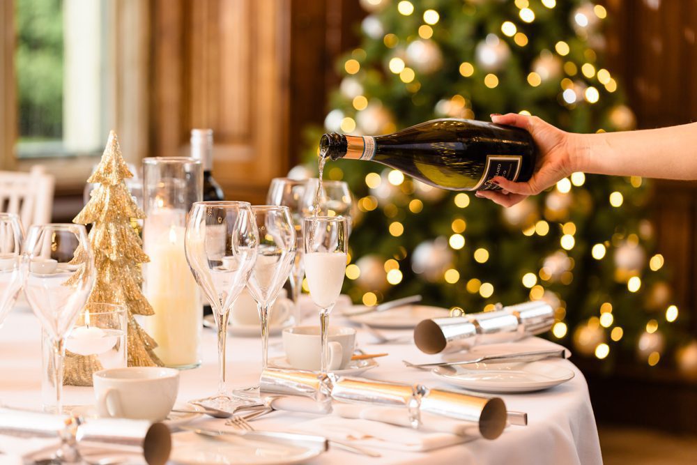 A hand pours champagne into a glass on a festive table, with a decorated Christmas tree in the background.