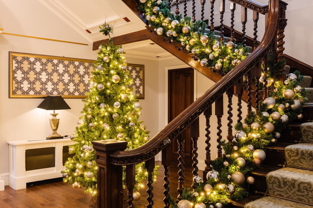 A decorated Christmas tree and garland with lights and ornaments on a staircase in a warmly lit hallway.