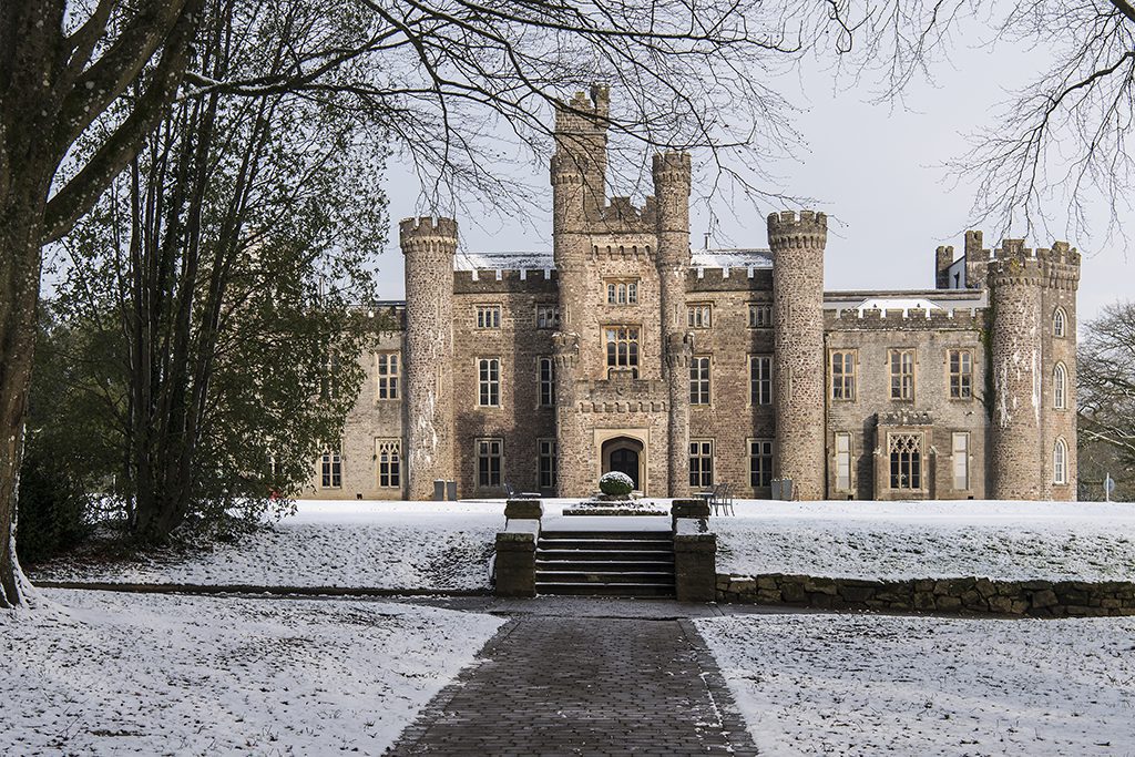 Stone castle with multiple towers and arched entrance, surrounded by trees and light snow on the ground.