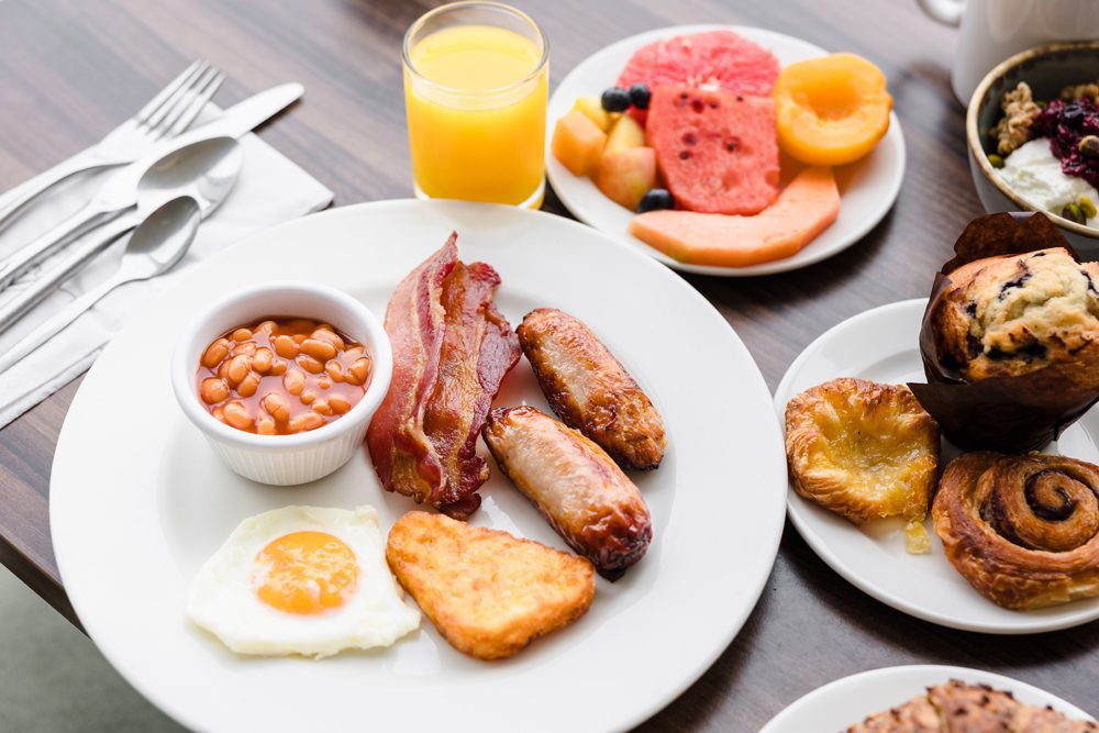 A breakfast plate with bacon, sausages, baked beans, hash brown, and fried egg, with fruit, juice, and pastries nearby.