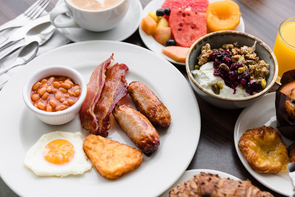 A breakfast plate with fried egg, hash brown, sausages, bacon, baked beans, yogurt with granola, fruit, juice, and coffee.