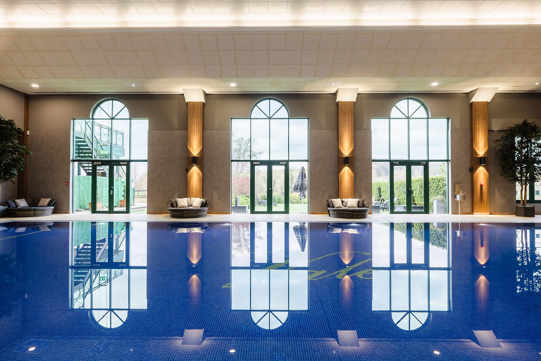 Indoor pool with blue water, large arched windows, lounge chairs, and views of greenery outside.