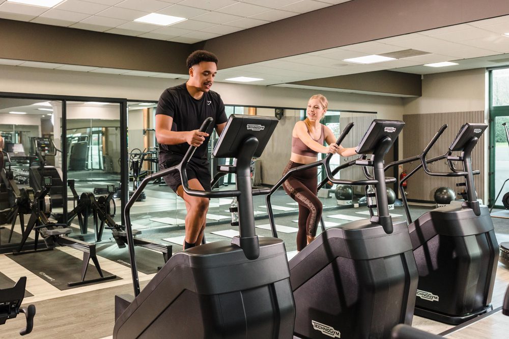 Two people exercise on stair machines in a gym with mirrors, weights, and fitness equipment in the background.