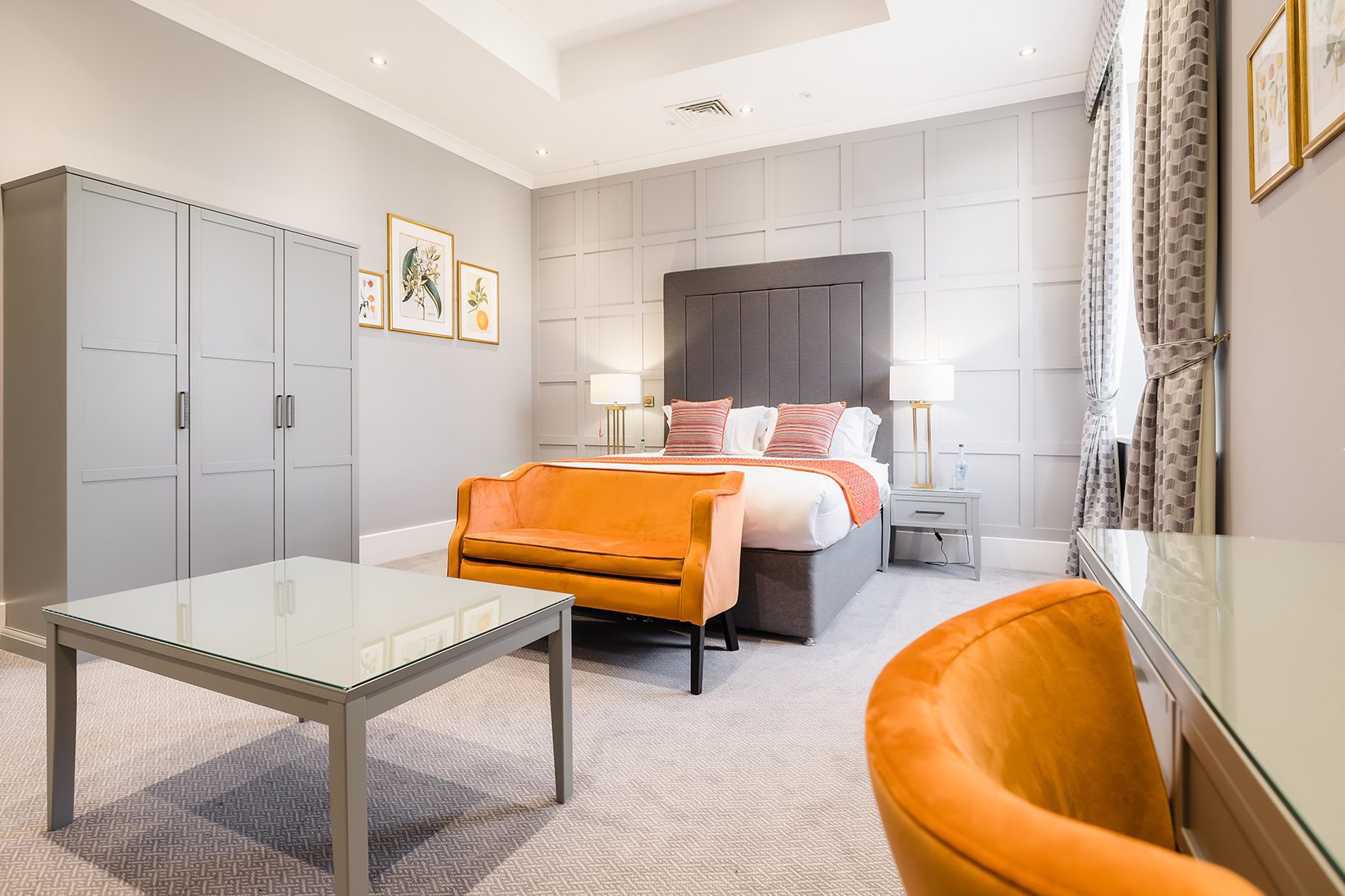 Modern bedroom with a gray wardrobe, bed, glass table, and orange velvet sofa and chair, with framed art on the walls.