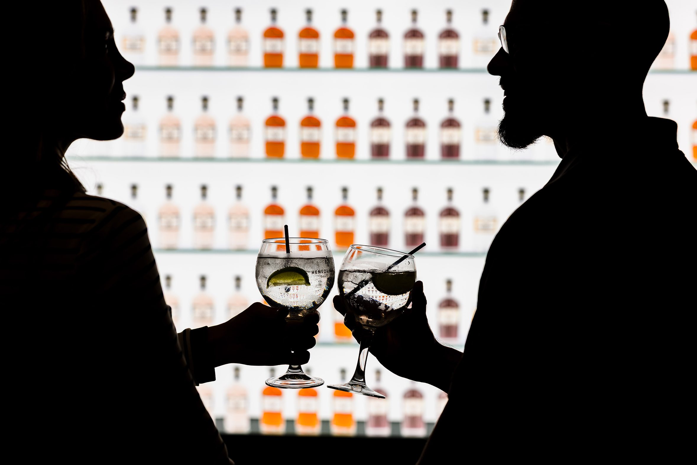 Silhouettes of two people clinking cocktails during stylish hotel breaks South Wales, bottles lined up on a bright shelf.