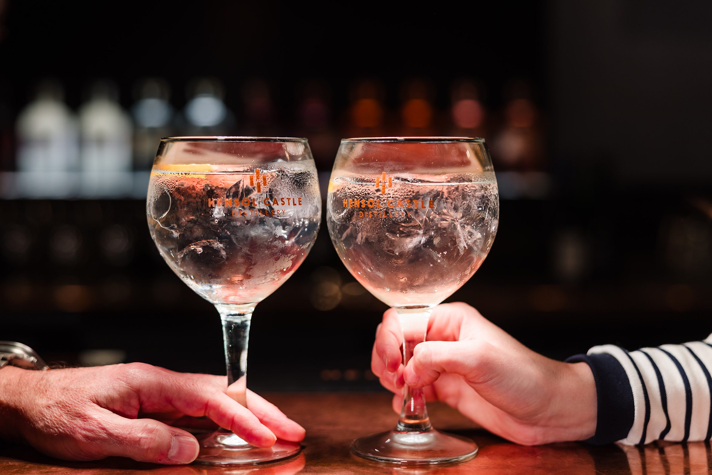 Two people toast gin and tonics at a bar, celebrating hotel room bookings with a blurred background.