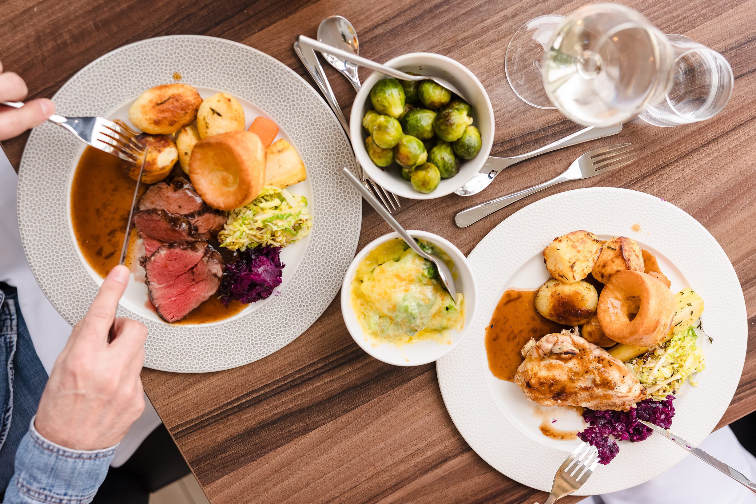 Two plates of Sunday lunch with meat, potatoes, Yorkshire pudding, veggies, Brussels sprouts, and drinks on a wooden table.