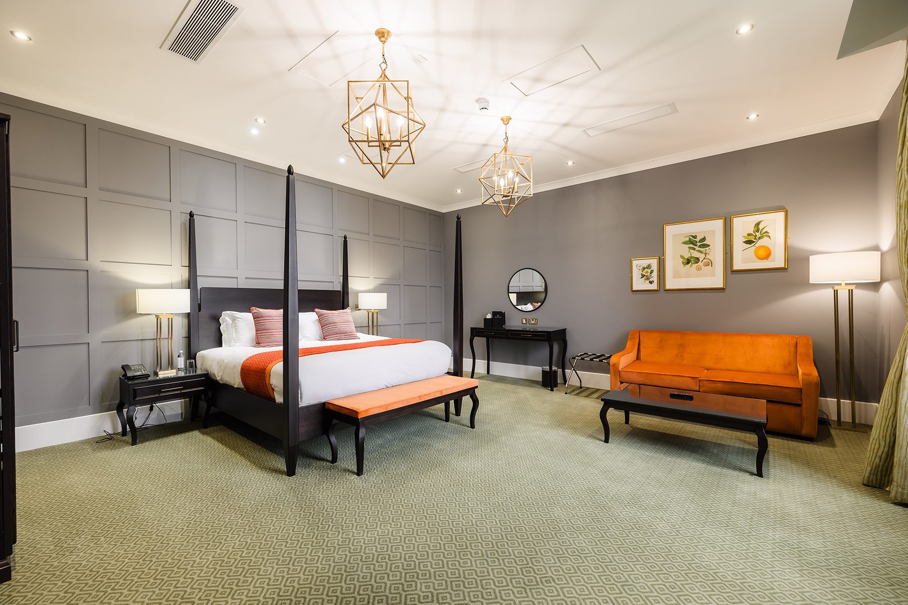 Spacious hotel room with a four-poster bed, orange bench, sofa, wall art, and geometric light fixtures.