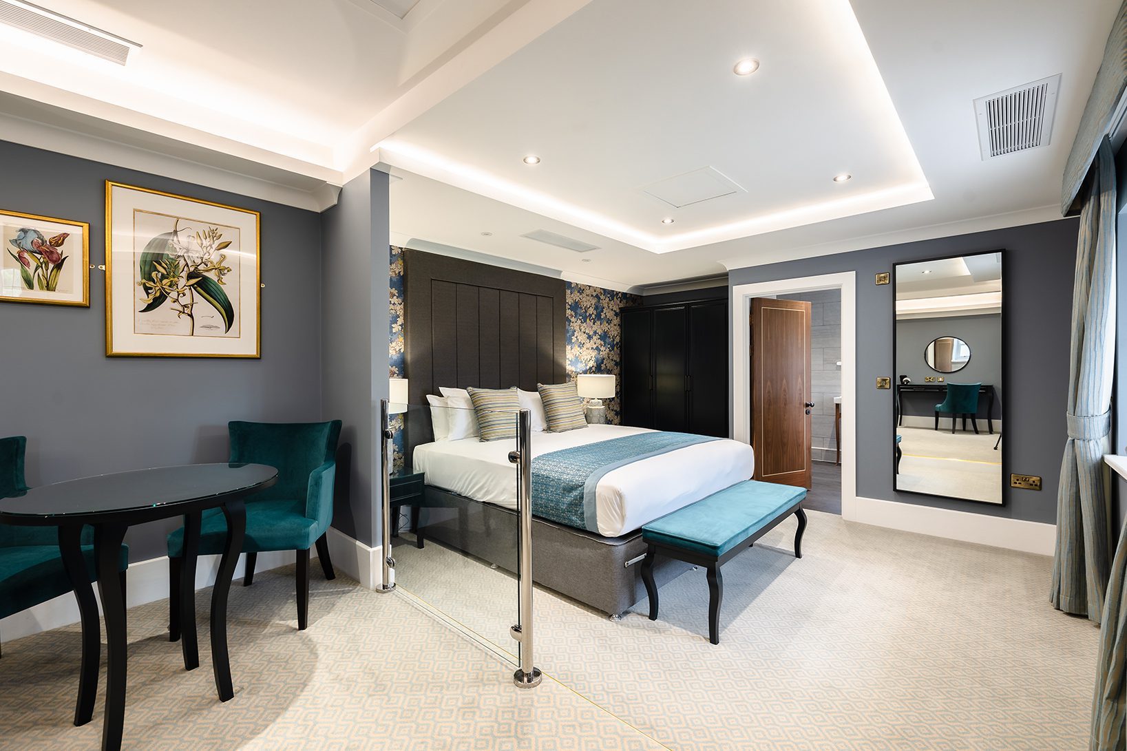 Modern hotel room with a double bed, teal seating area, large mirror, and floral artwork on the wall.
