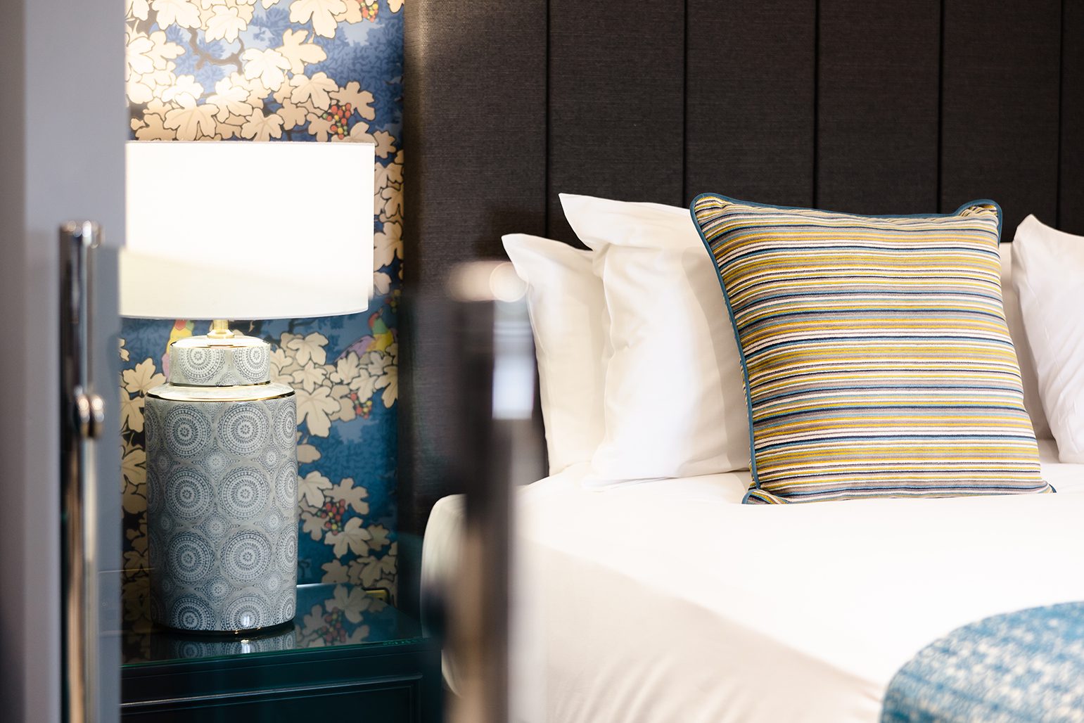 A bed with a striped pillow next to a nightstand with a lamp and blue-patterned wallpaper in the background.