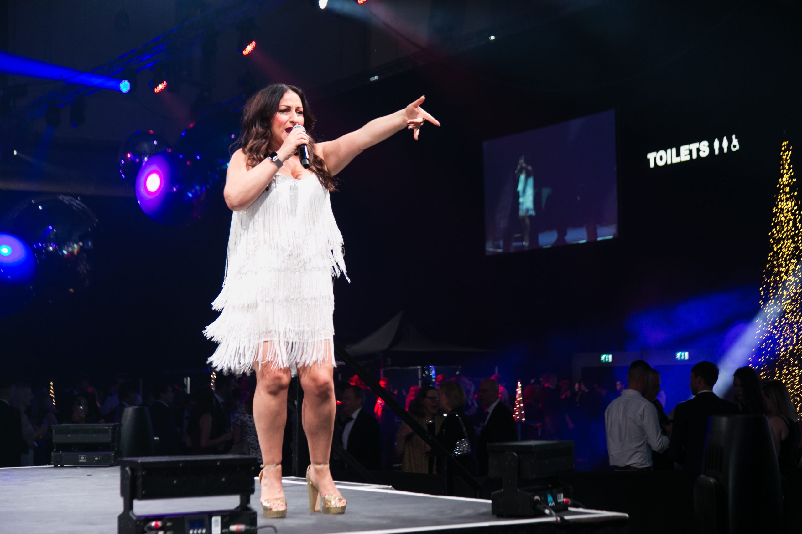 A woman in a white fringed dress sings and points on stage at an indoor event with an audience and lights.