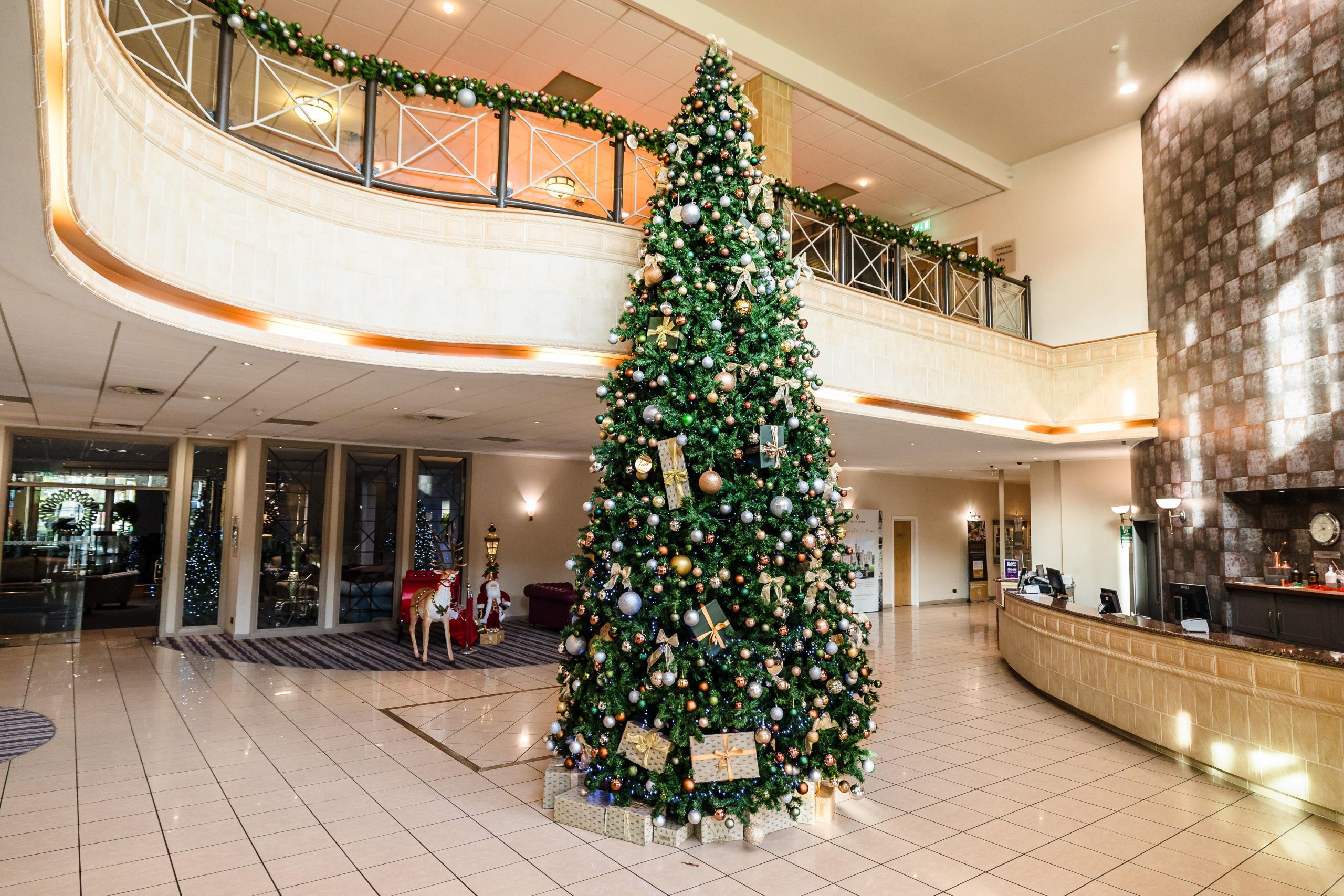 A large decorated Christmas tree stands in the lobby of a spacious, modern building with tiled floors and balcony railings.