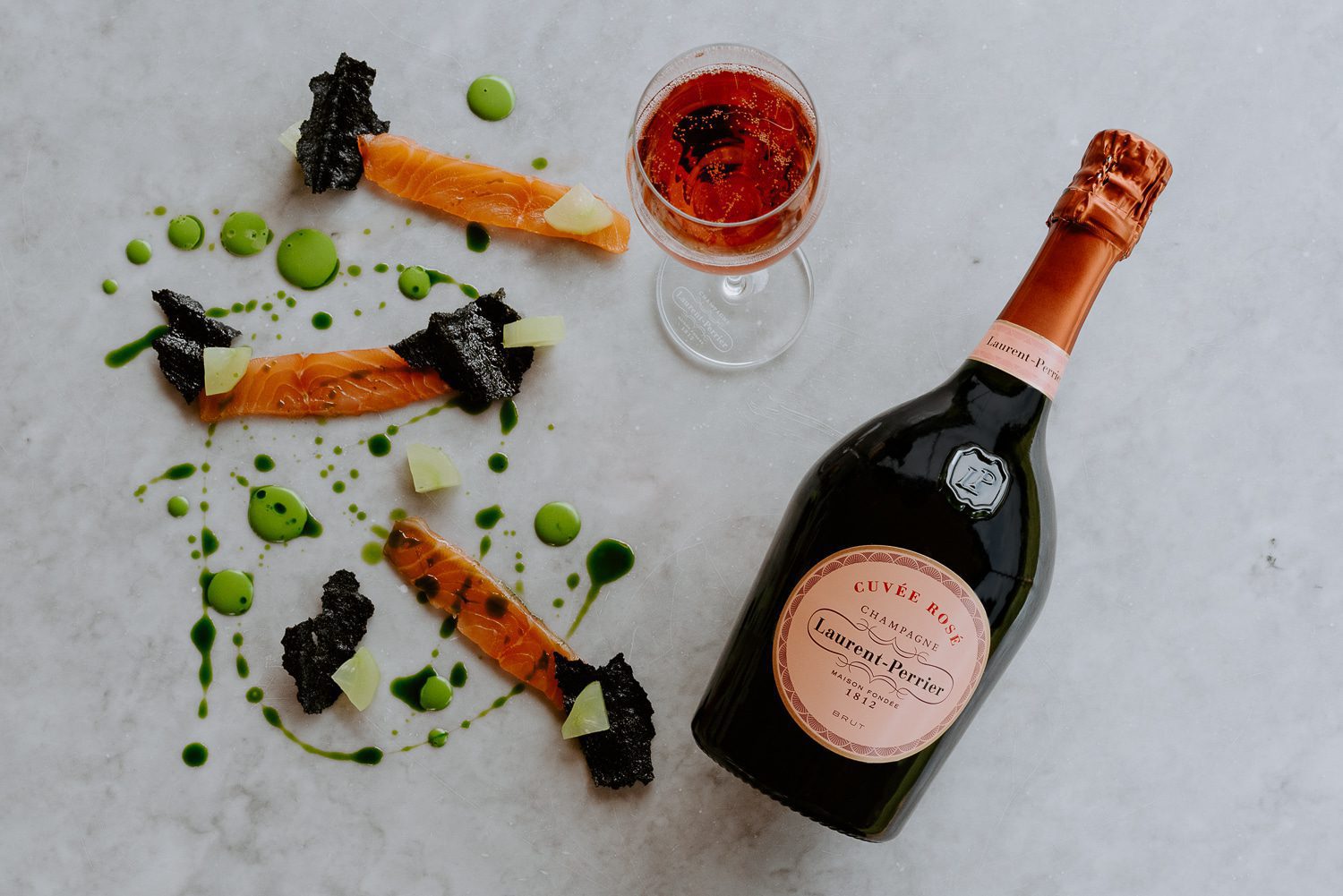 Bottle of Laurent-Perrier Cuvée Rosé, glass of rosé wine, and plated salmon with sauce and garnishes on a marble surface.