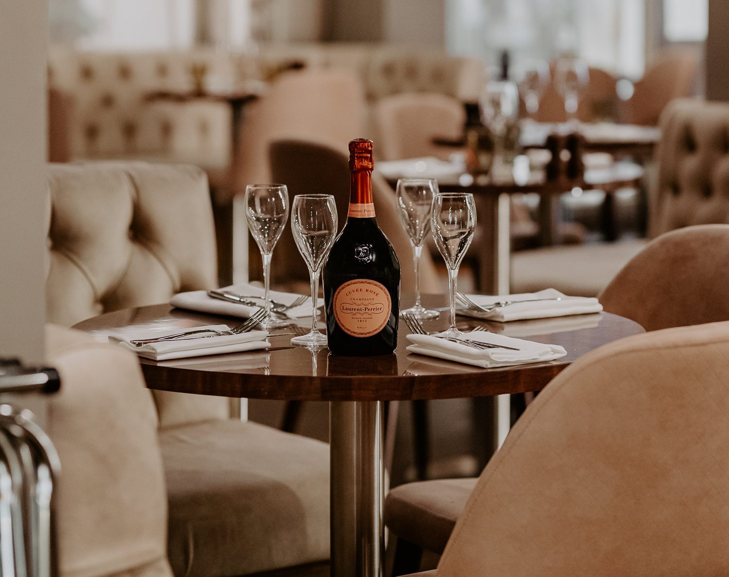A round dining table set with a bottle of champagne, four glasses, serviettes, and cutlery in an elegant restaurant.