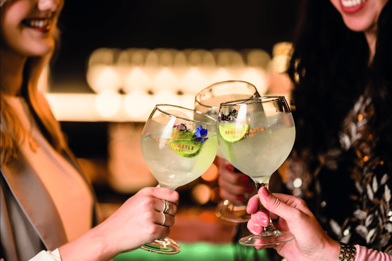 Three people raise large gin glasses in a stylish lounge after making seamless hotel room reservations.