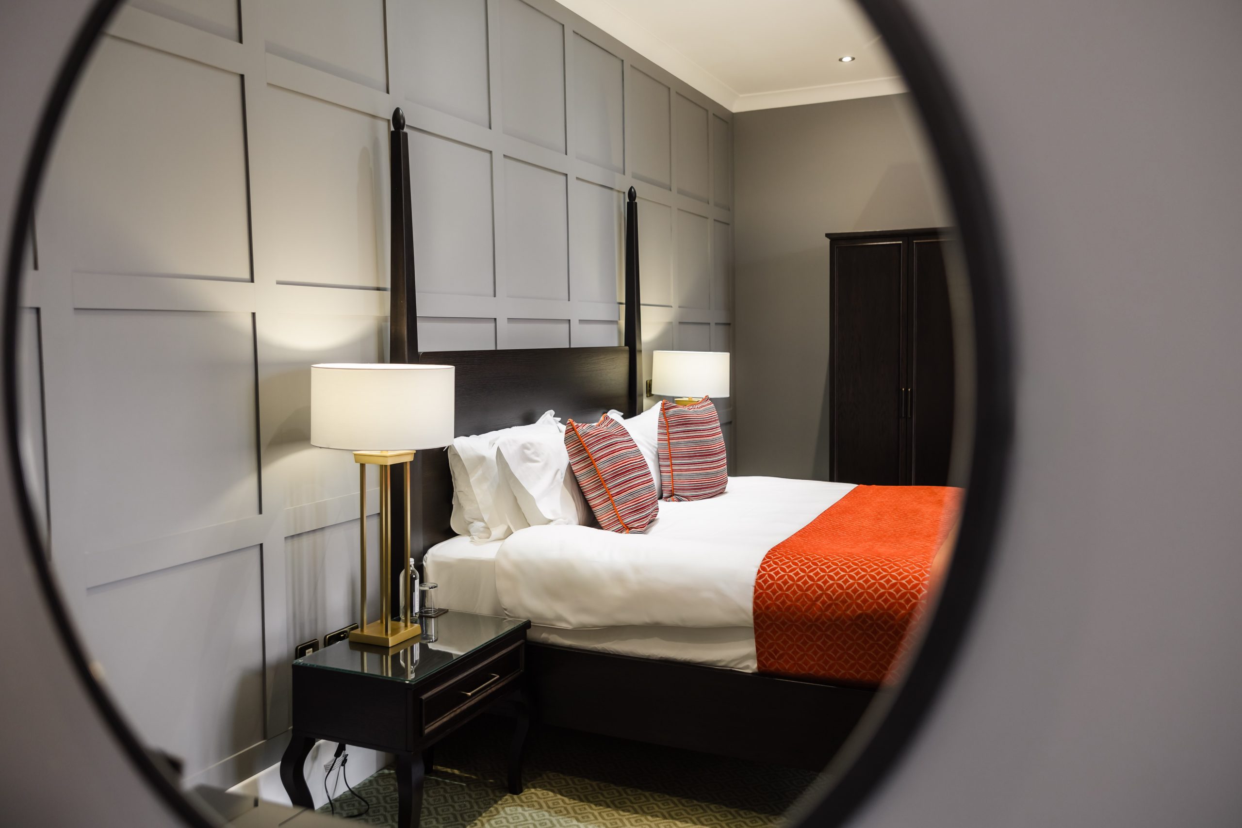 A neatly made bed with orange accents, flanked by lamps, seen through a circular mirror in a modern hotel room.