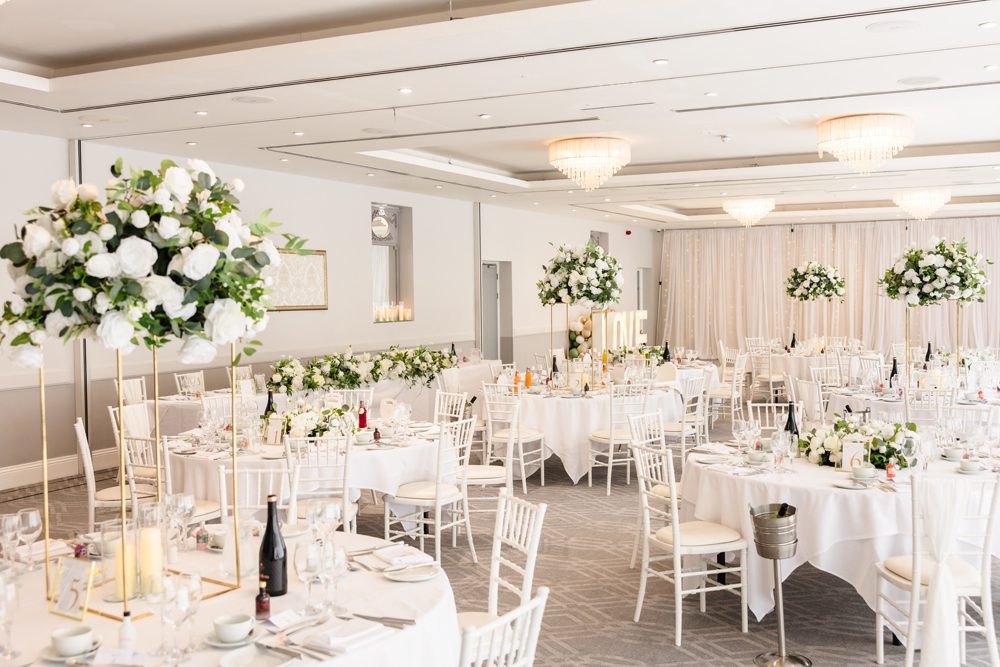 A bright, elegant banqueting hall set for a September wedding with round tables, white chairs, and large floral centrepieces.